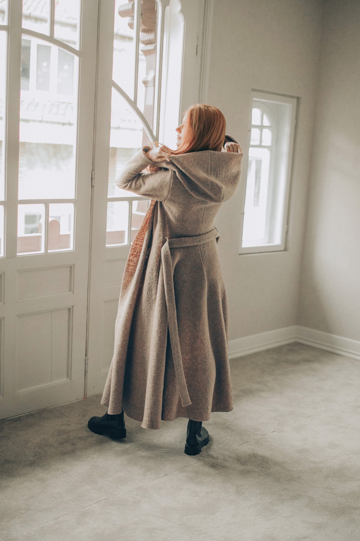 Woman in a long beige coat standing in a room with large windows.