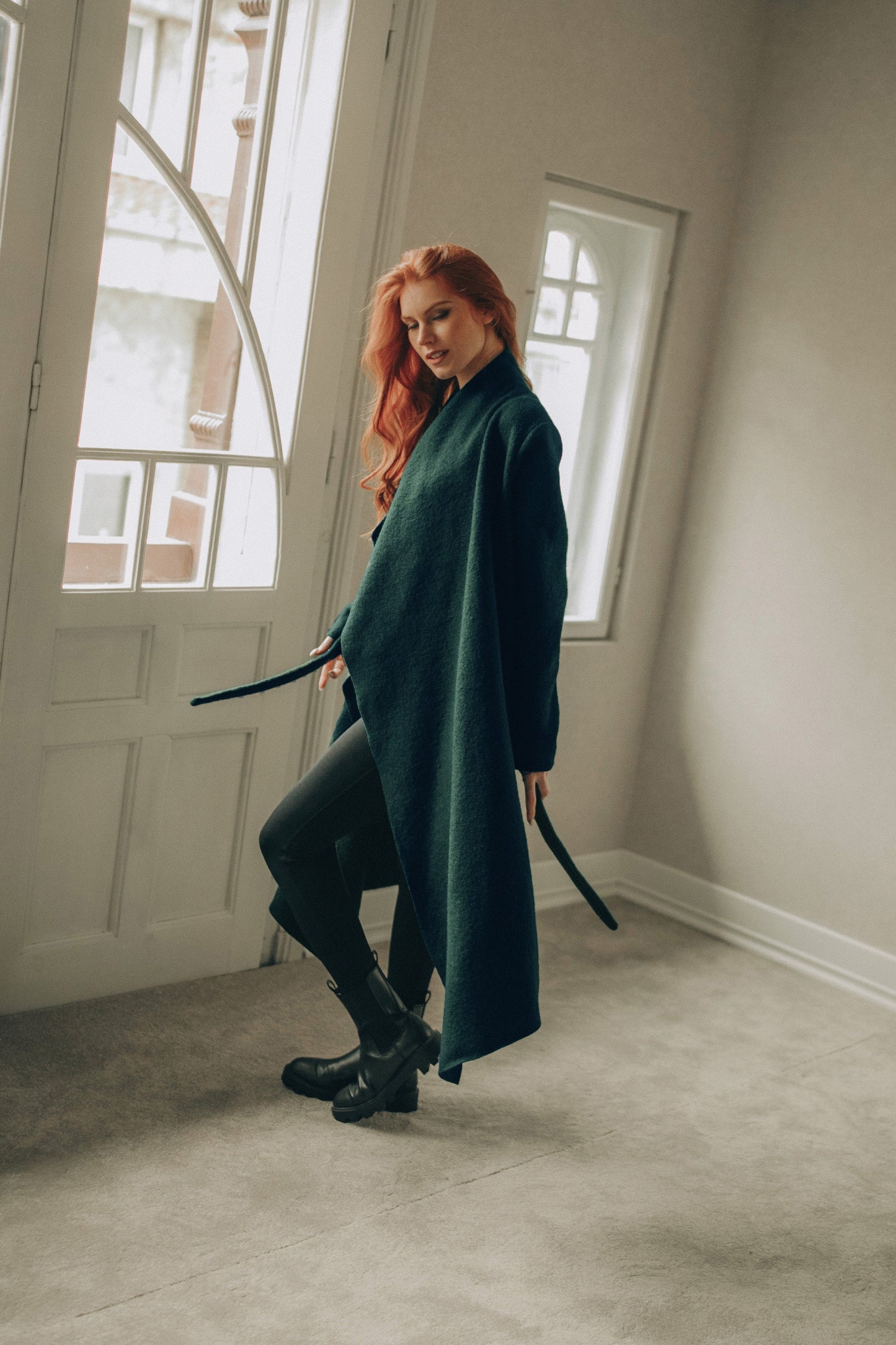 Woman in a dark green coat standing in a room with large windows.