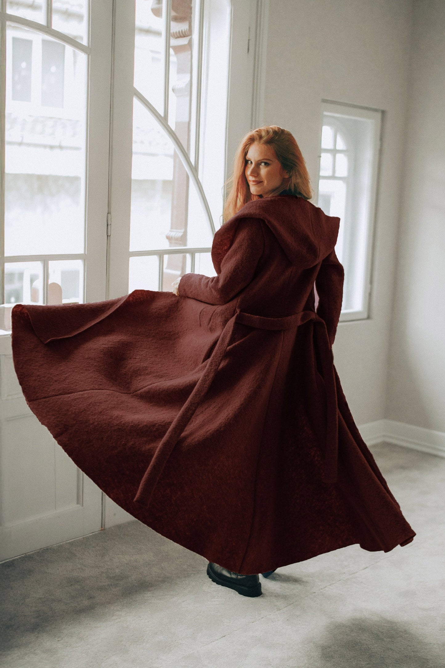 Woman wearing a flowing burgundy maxi wool hooded cardigan coat with an adjustable belt and hood indoors