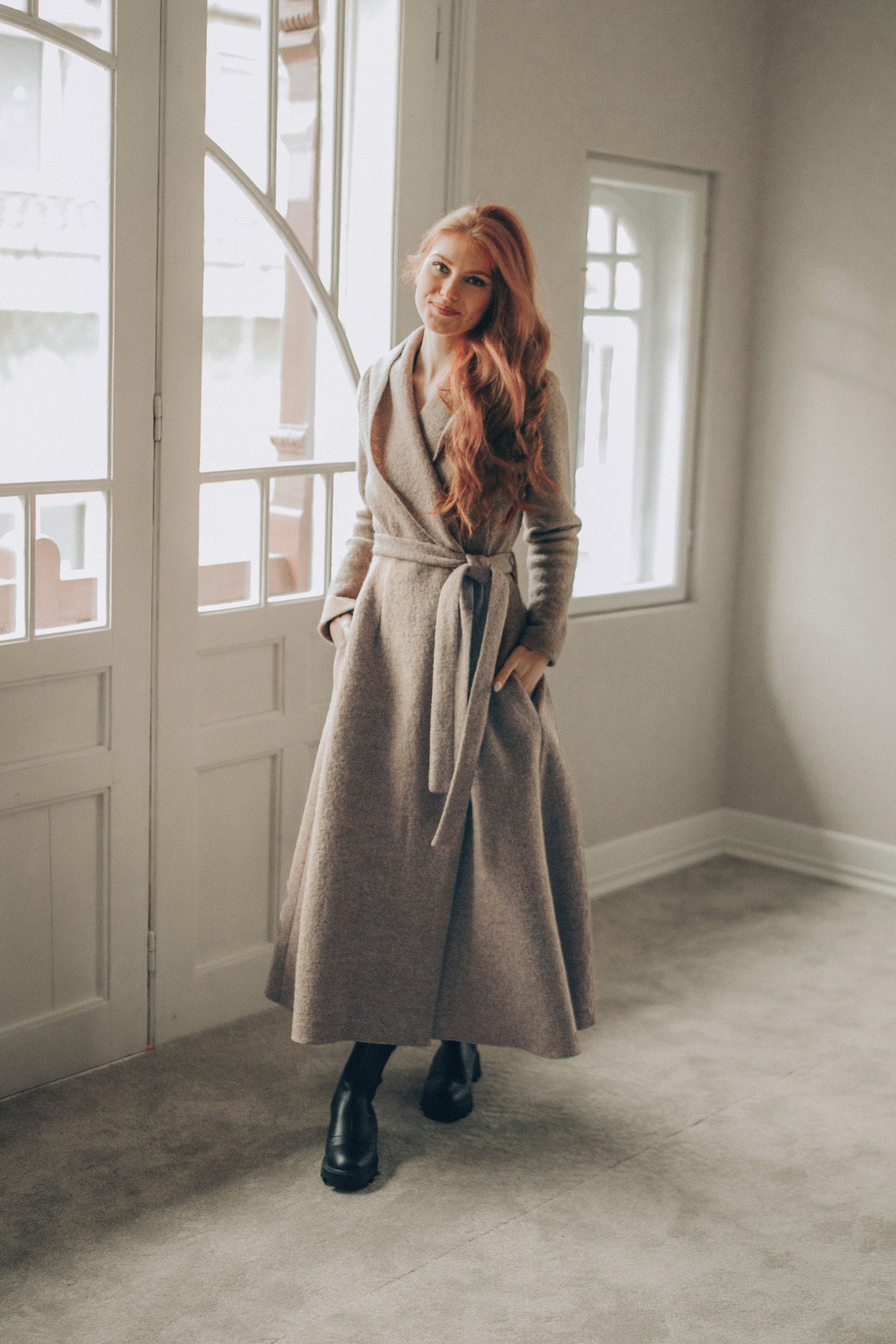 Woman in a long coat standing in a room with large windows.