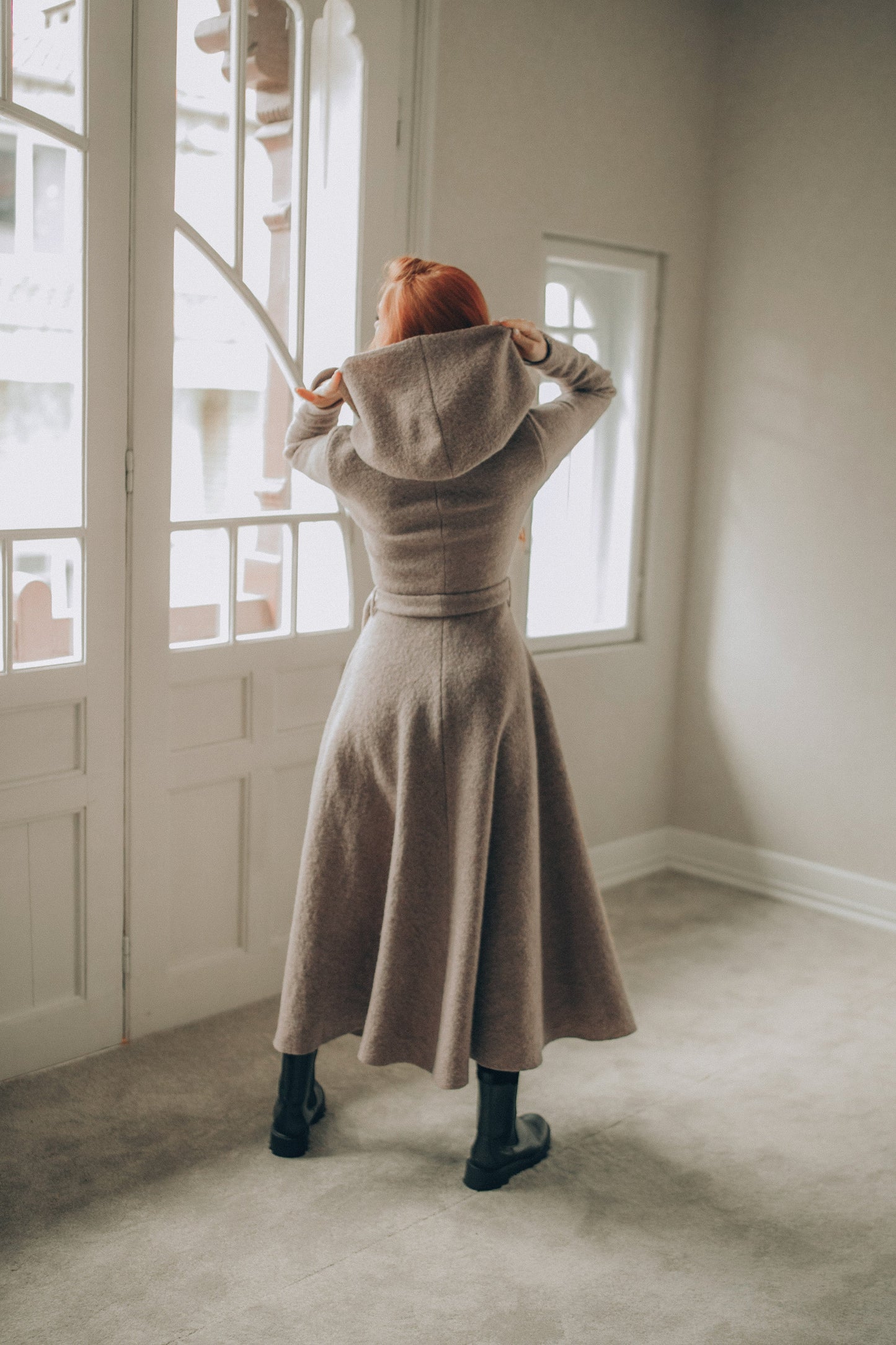 Woman wearing a long beige coat standing in a room with large windows.