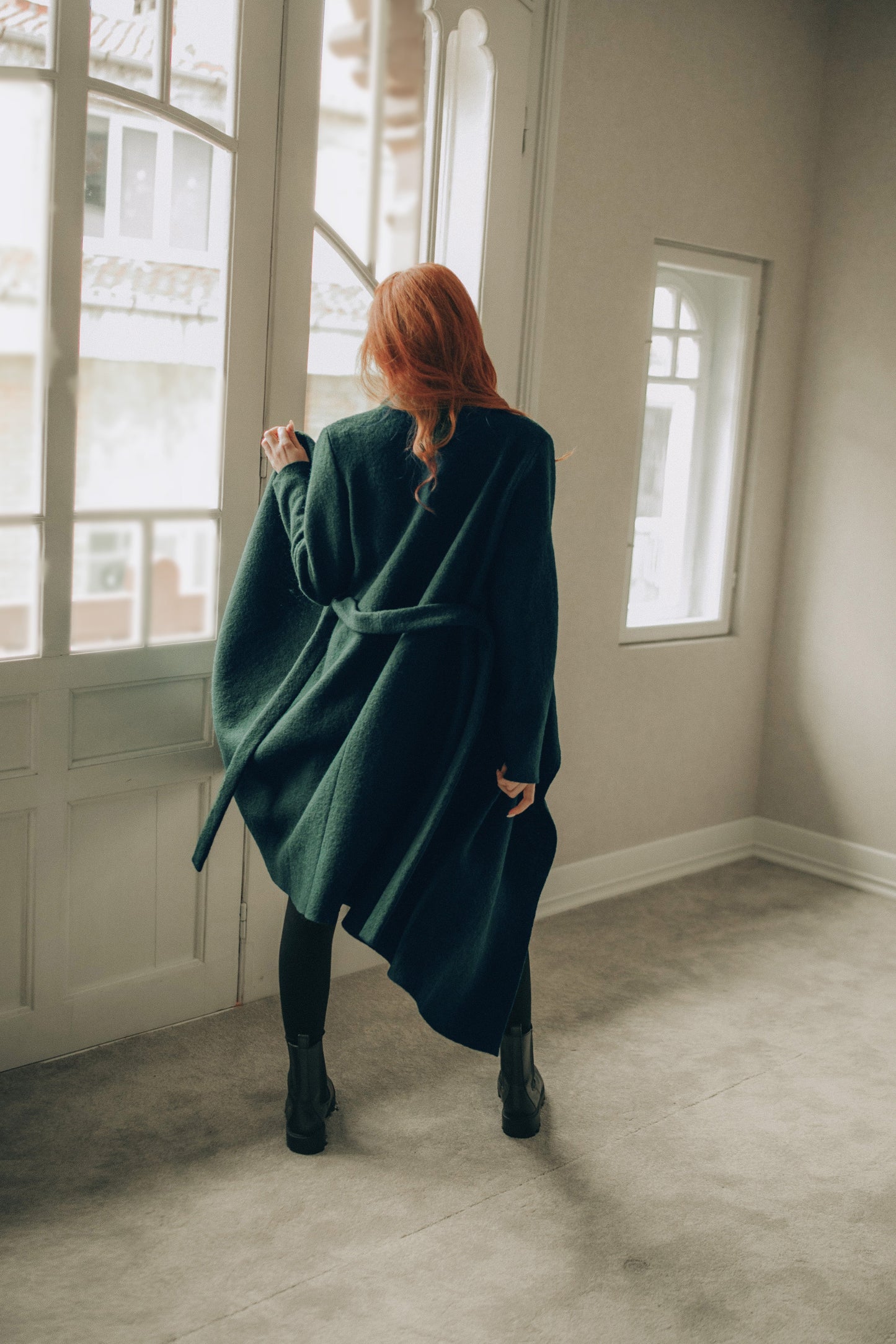 Woman in a green coat standing in a room with large windows.