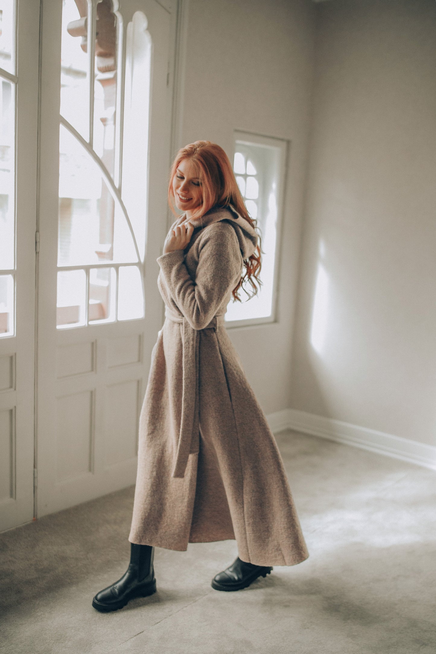 Woman in a long beige coat standing in a room with large windows.