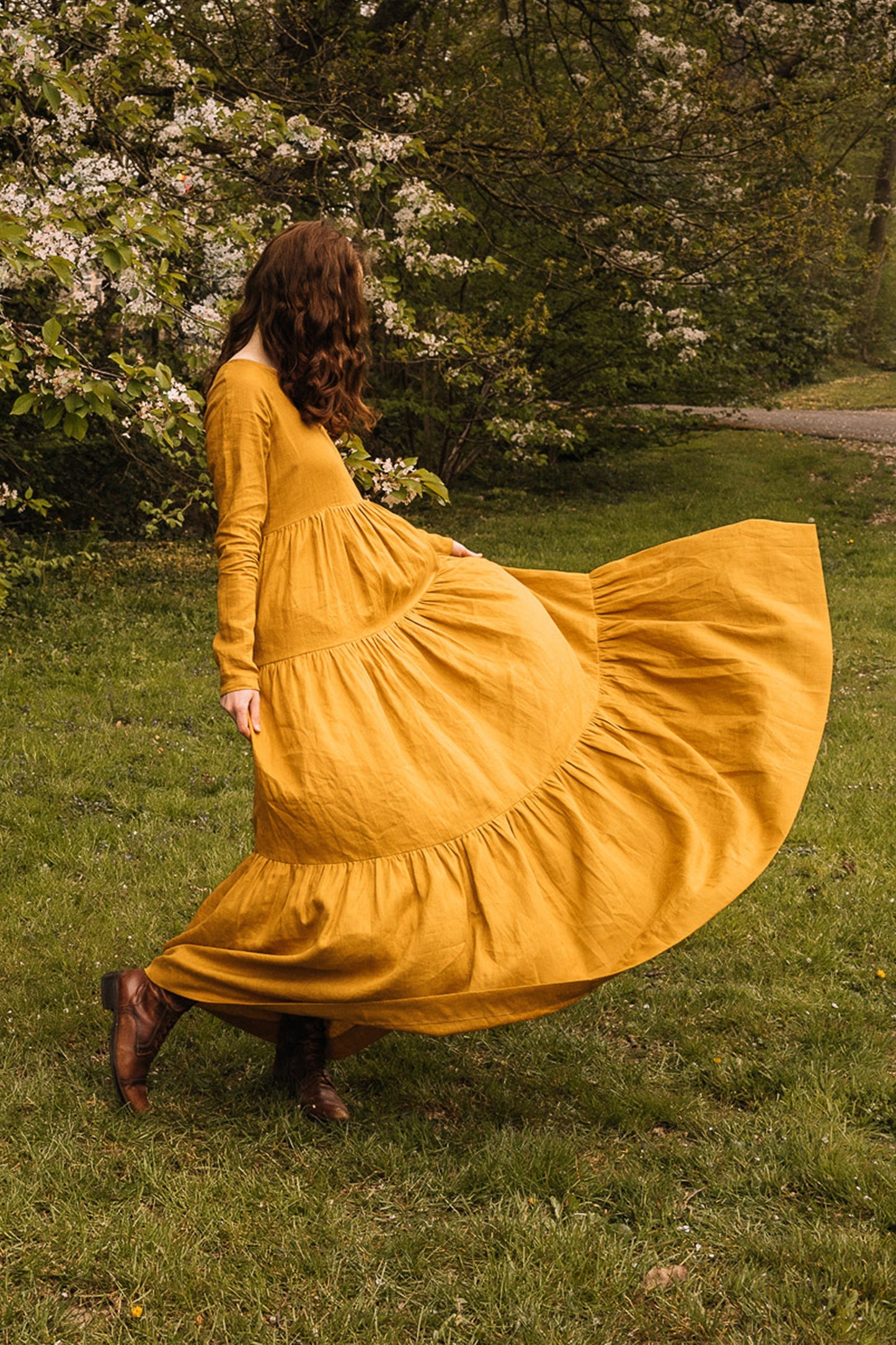 Woman wearing a mustard bohemian linen dress twirling outdoors with greenery and blossoms in background