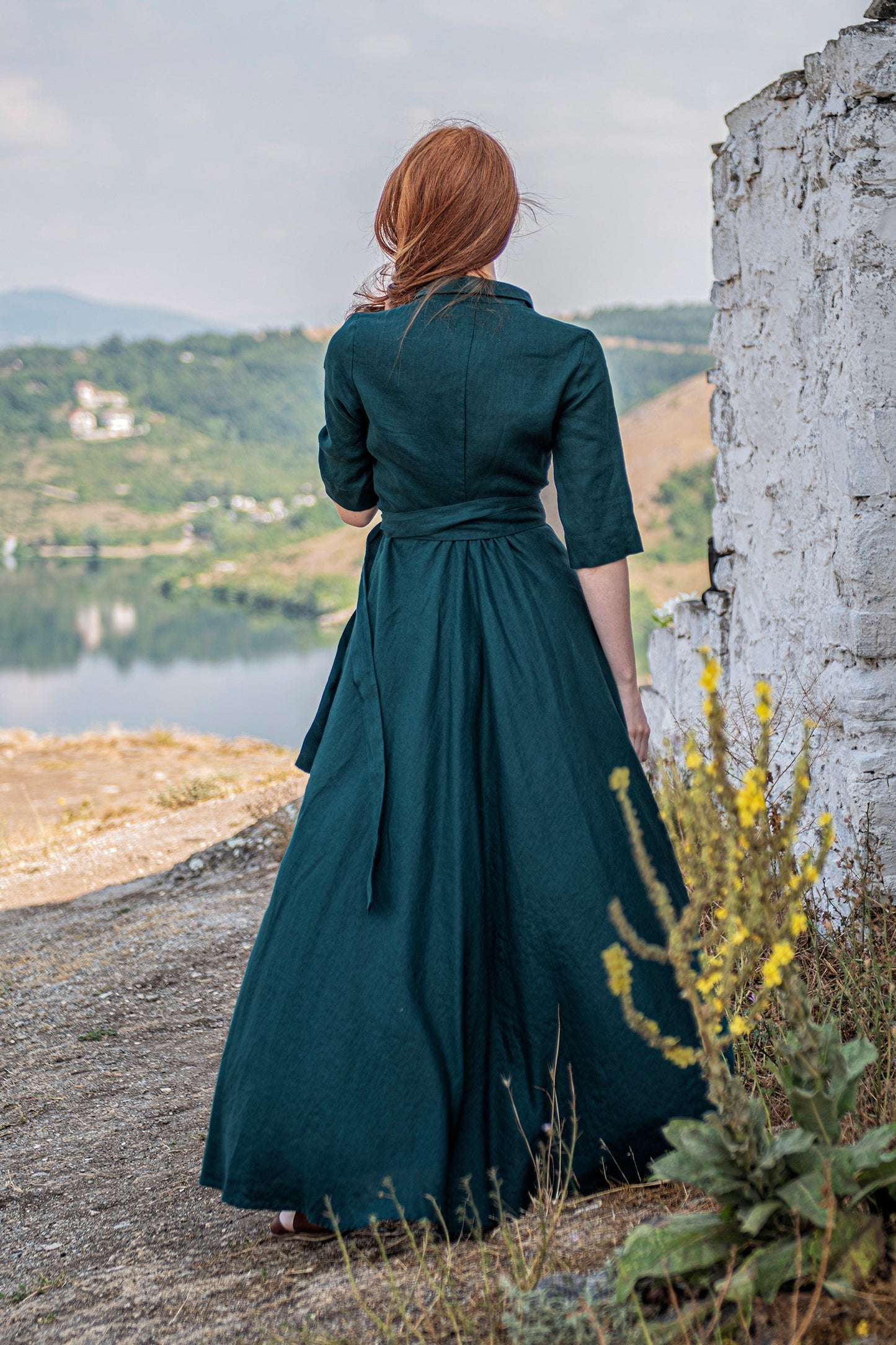 Woman wearing dark green linen maxi dress with tie waist standing outdoors near a stone wall