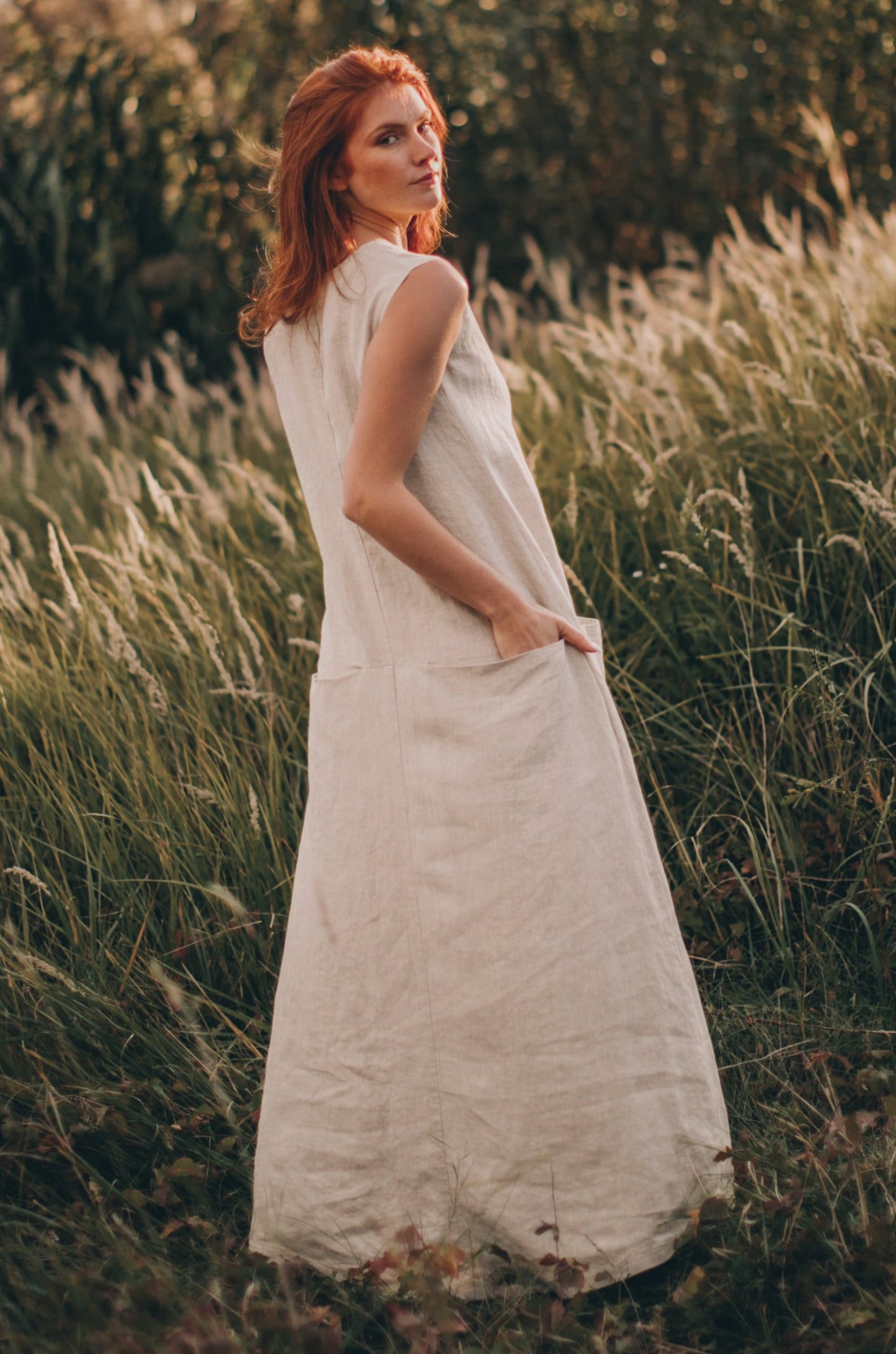Woman wearing a sleeveless beige linen maxi dress with pockets standing in a grassy field
