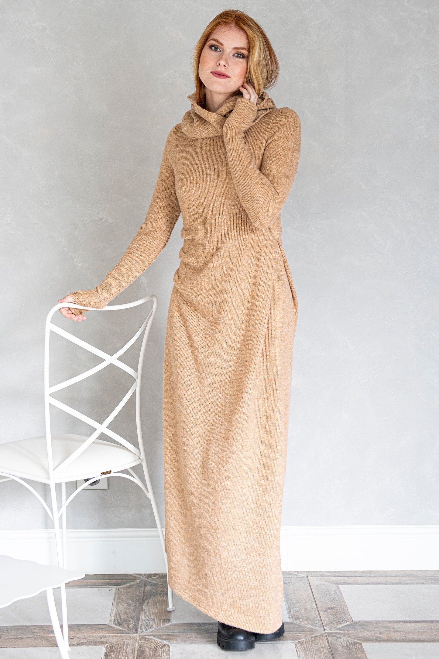 Woman wearing beige hooded knit dress with side gathered accent standing by white chair