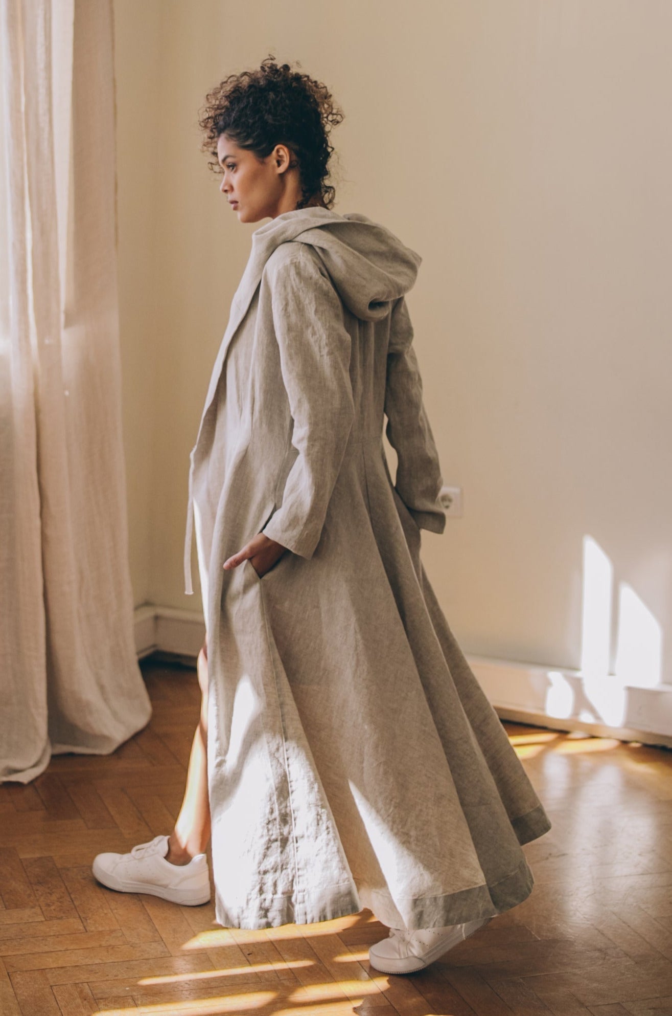 Woman wearing elegant heavyweight summer linen coat with hood and tie closure standing indoors