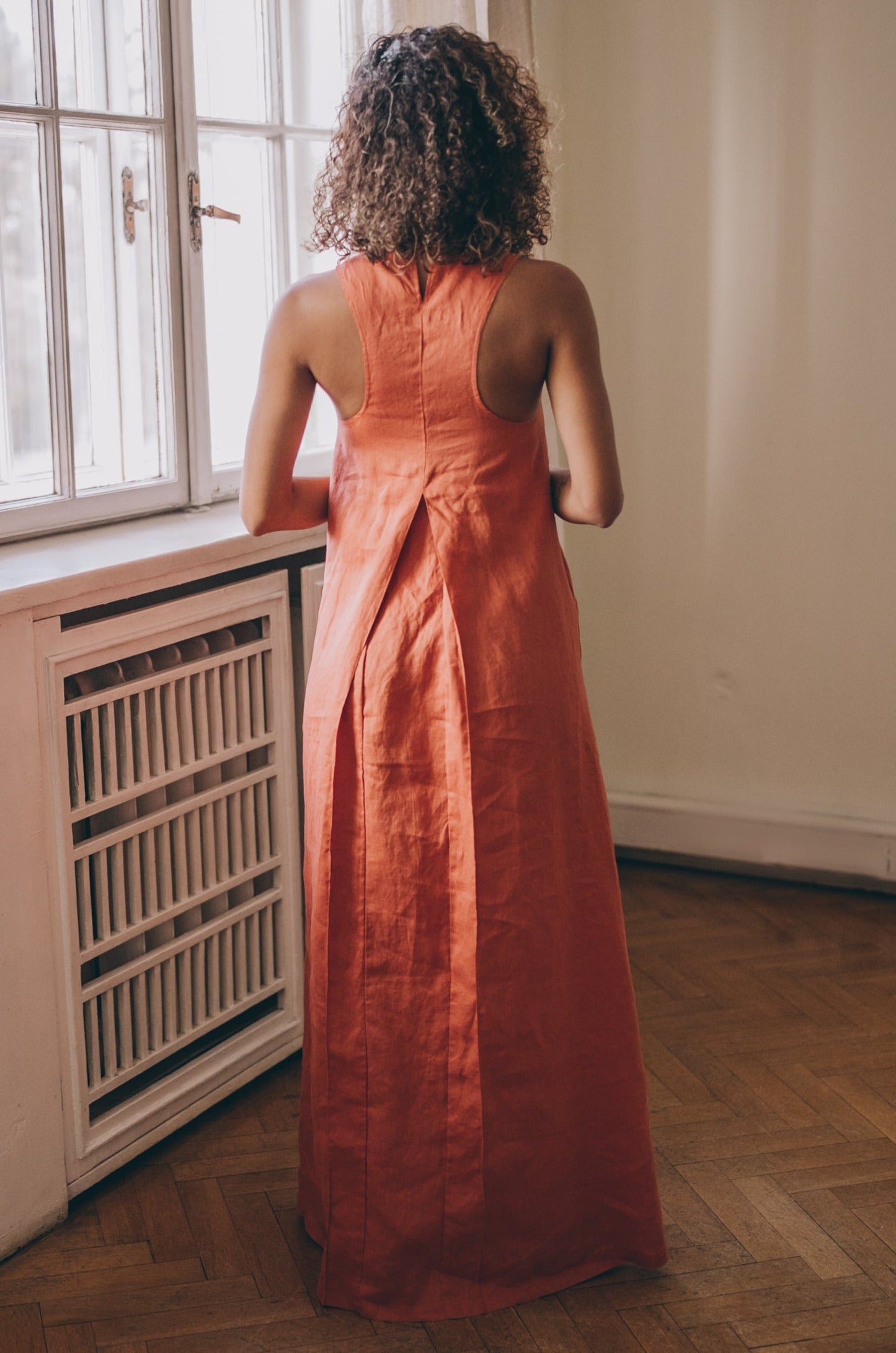 Woman wearing a sleeveless minimalist linen dress in rusty orange with back pleat detail standing by a window