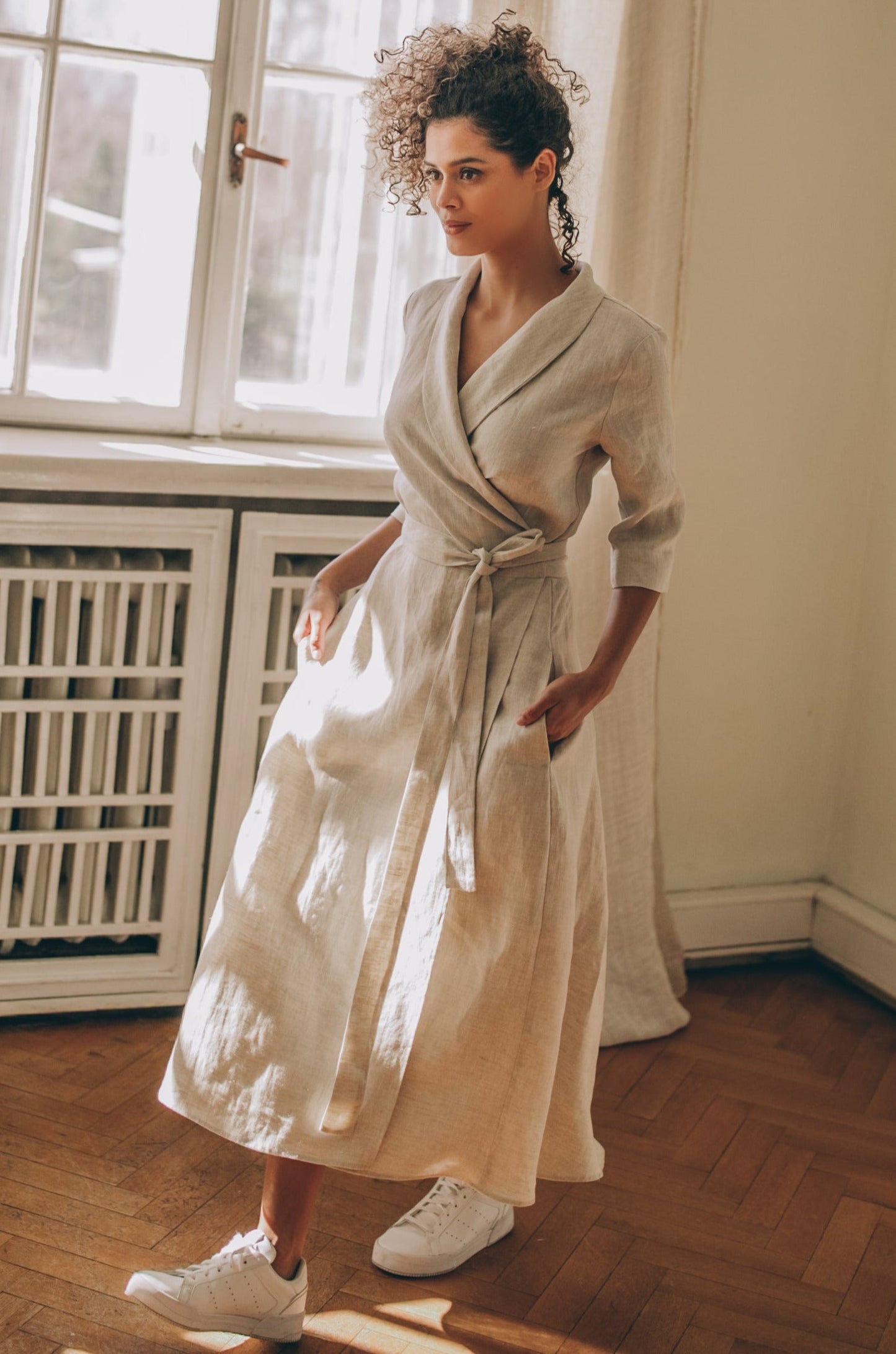 Woman wearing a beige midi length linen dress with tie waist and V-neck collar indoors