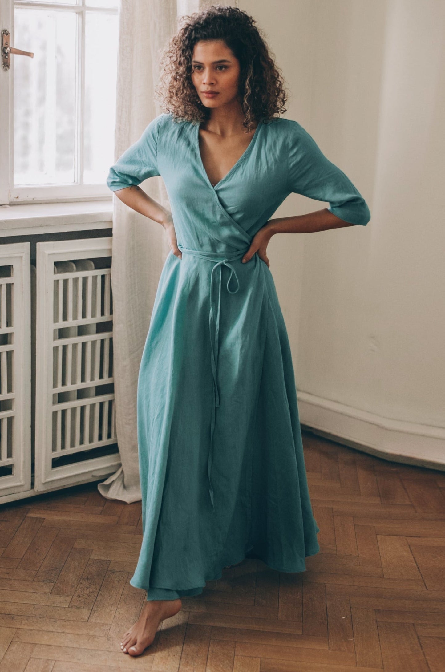 Woman wearing blue green linen maxi dress with tie waist and 3/4 sleeves indoors