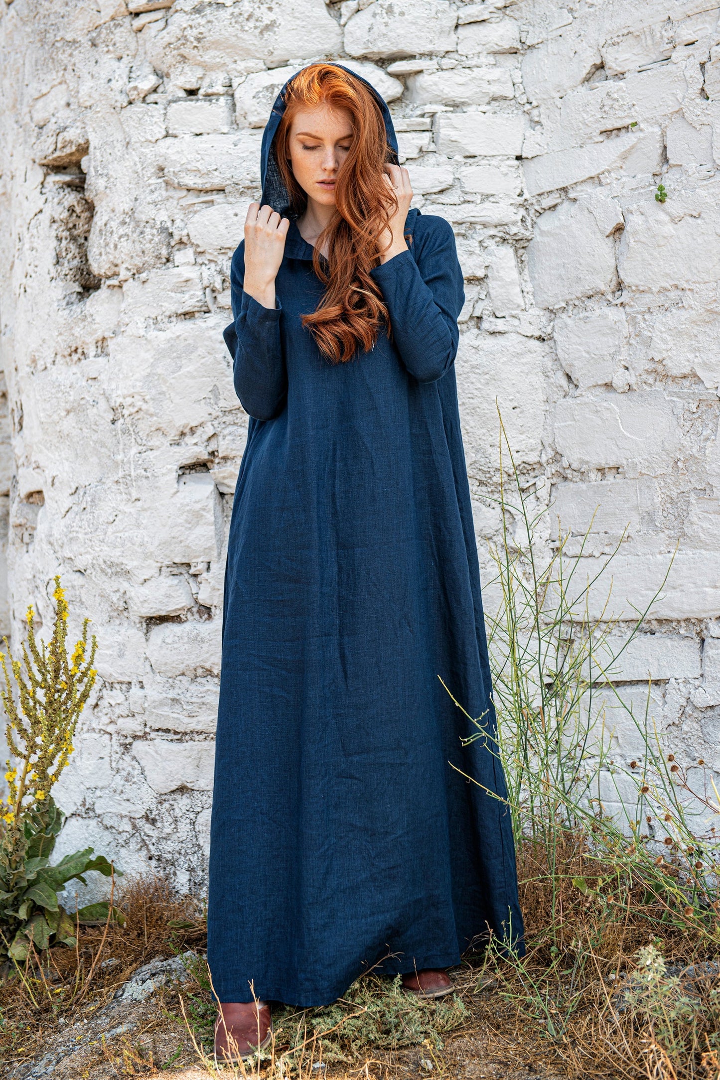 Woman wearing a long sleeve hooded linen dress standing outdoors against a stone wall.