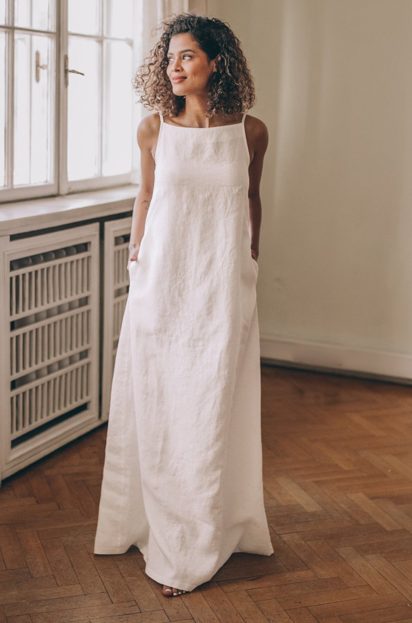 Woman wearing sleeveless linen maxi dress with boat neckline and pockets standing indoors
