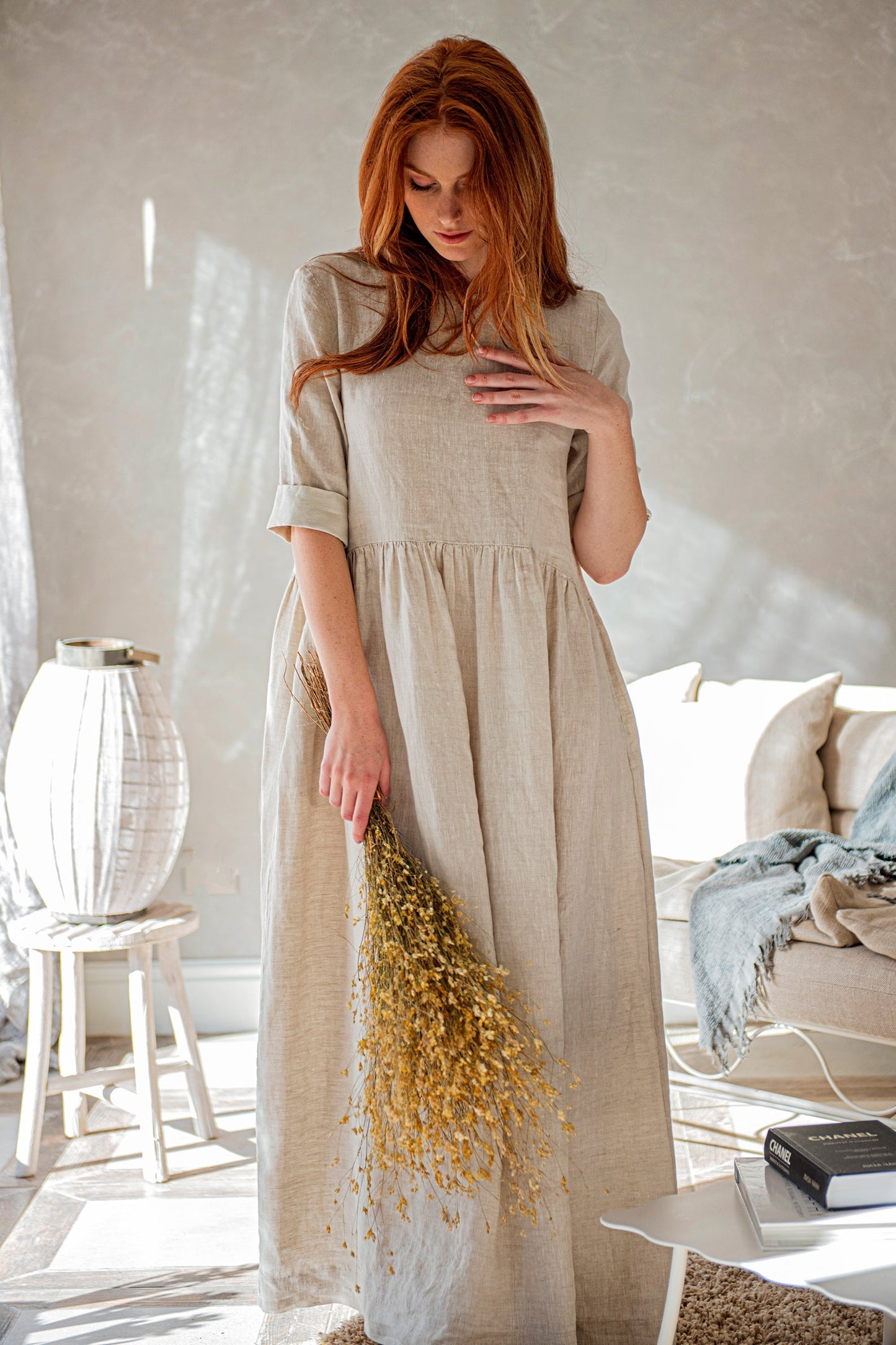 Woman wearing natural shade linen smock dress with elbow sleeves and pleated bodice holding dried flowers indoors