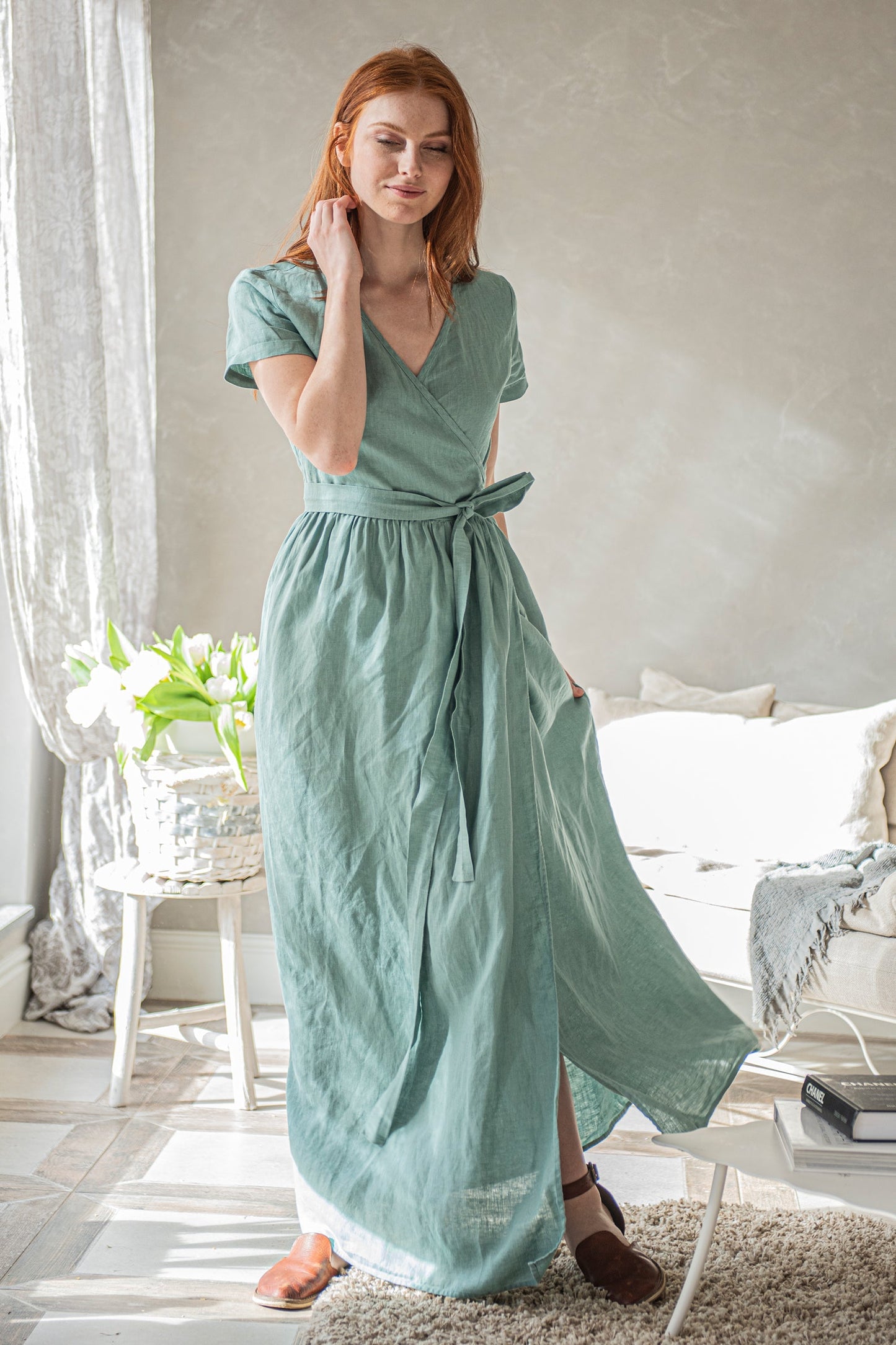 Woman wearing a soft green long linen wrap dress with short sleeves and tie waist in a bright room