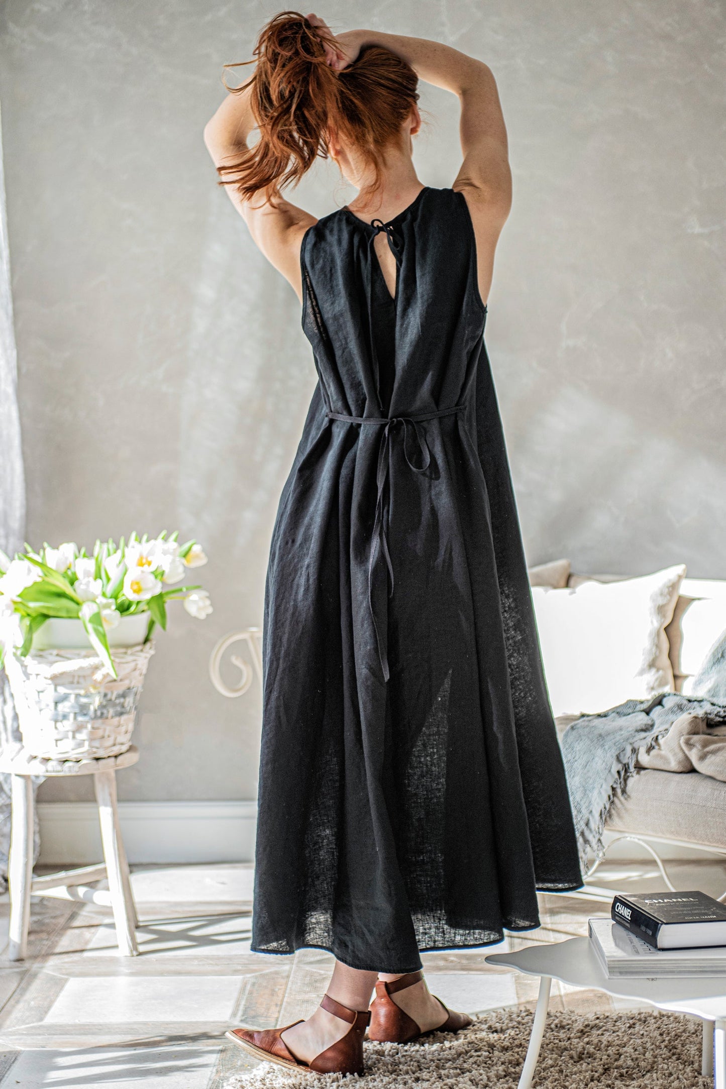 Woman wearing black sleeveless linen loose dress with tie knot closure at the back in bright room