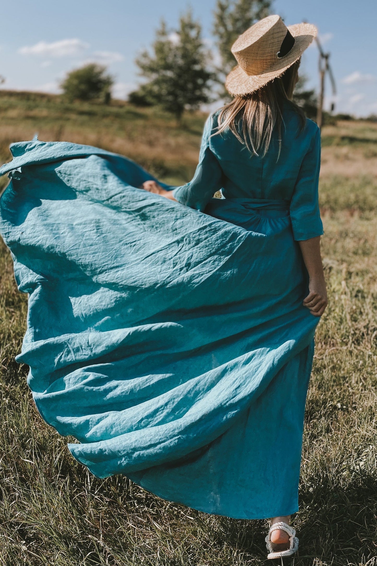 Woman wearing a flowing blue-green long linen summer dress with a tie belt and a straw hat standing in a field