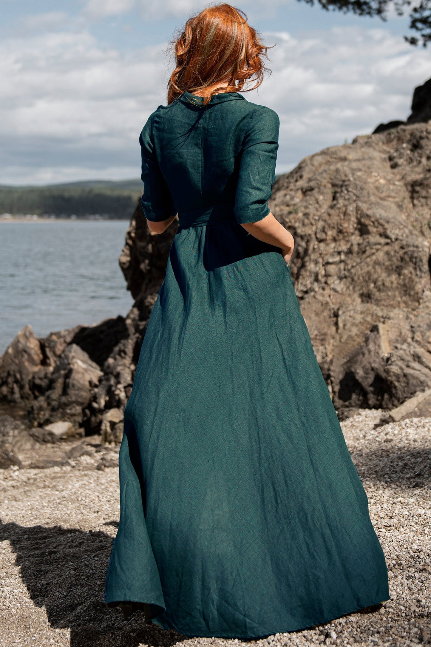 Woman wearing green linen wrap dress with tie waist standing near rocky shore