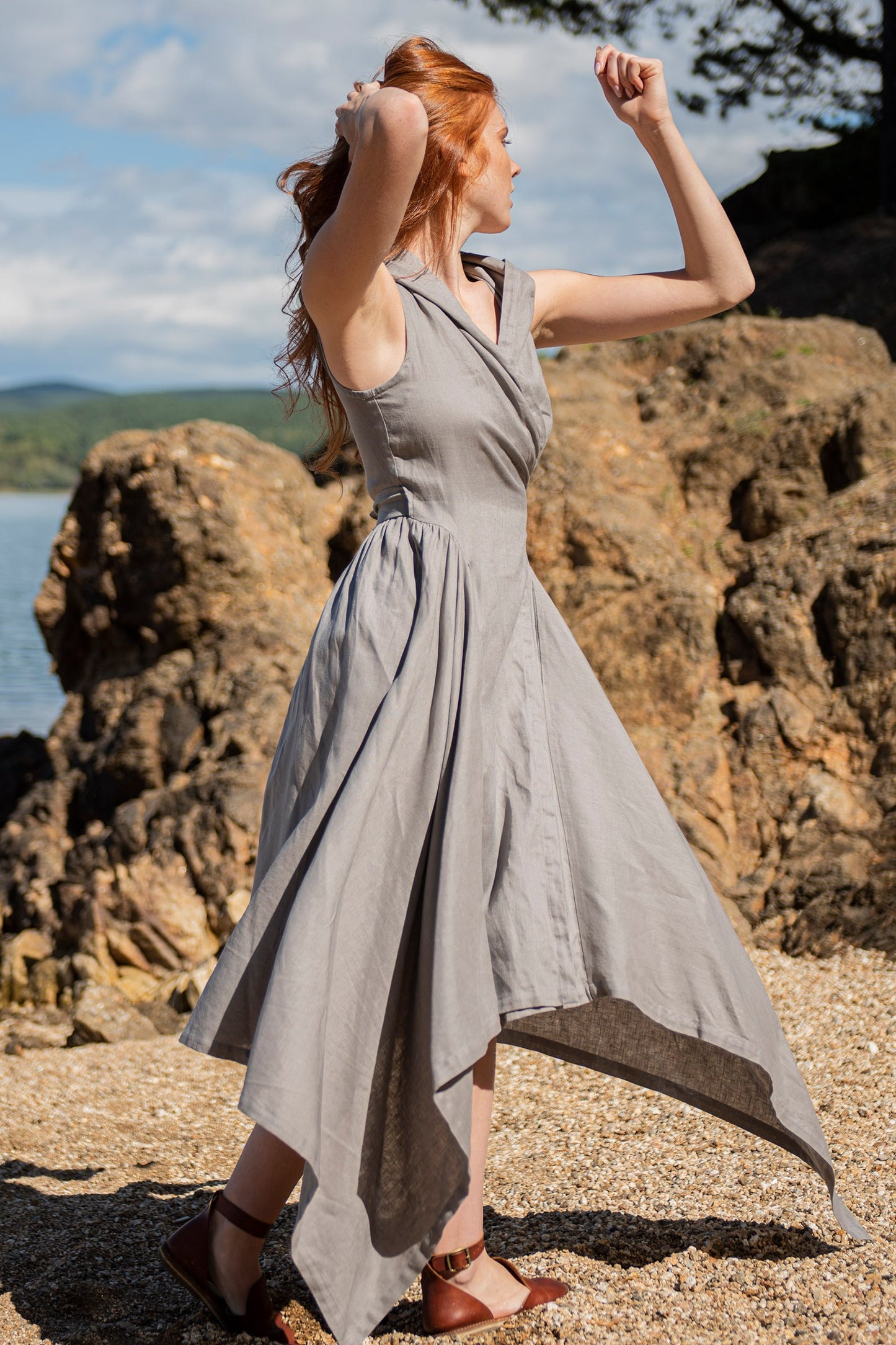 Woman wearing natural gray hooded linen asymmetric dress with tie belt on rocky beach