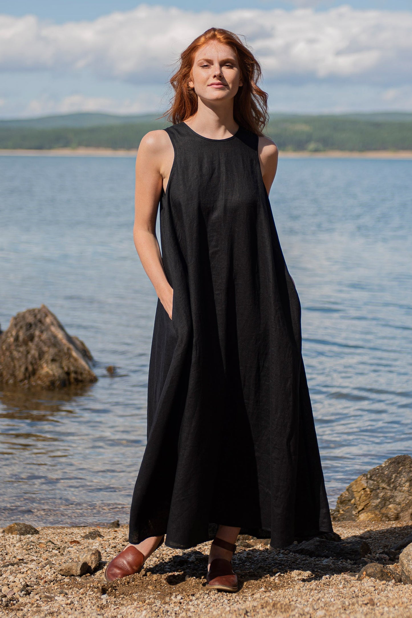 Woman wearing black sleeveless classic linen maxi dress with side pockets standing by water at the beach