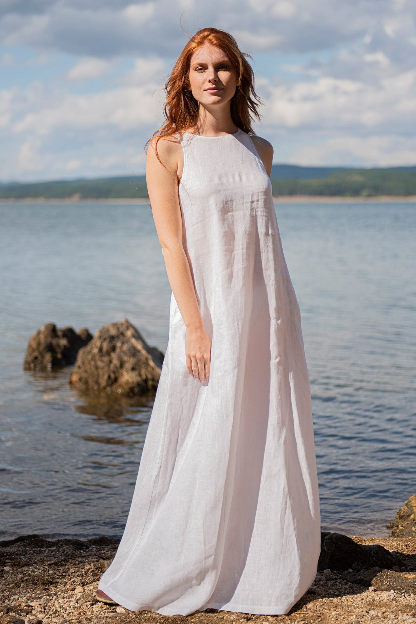 Woman wearing a sleeveless white minimalist linen dress standing by the rocky beach