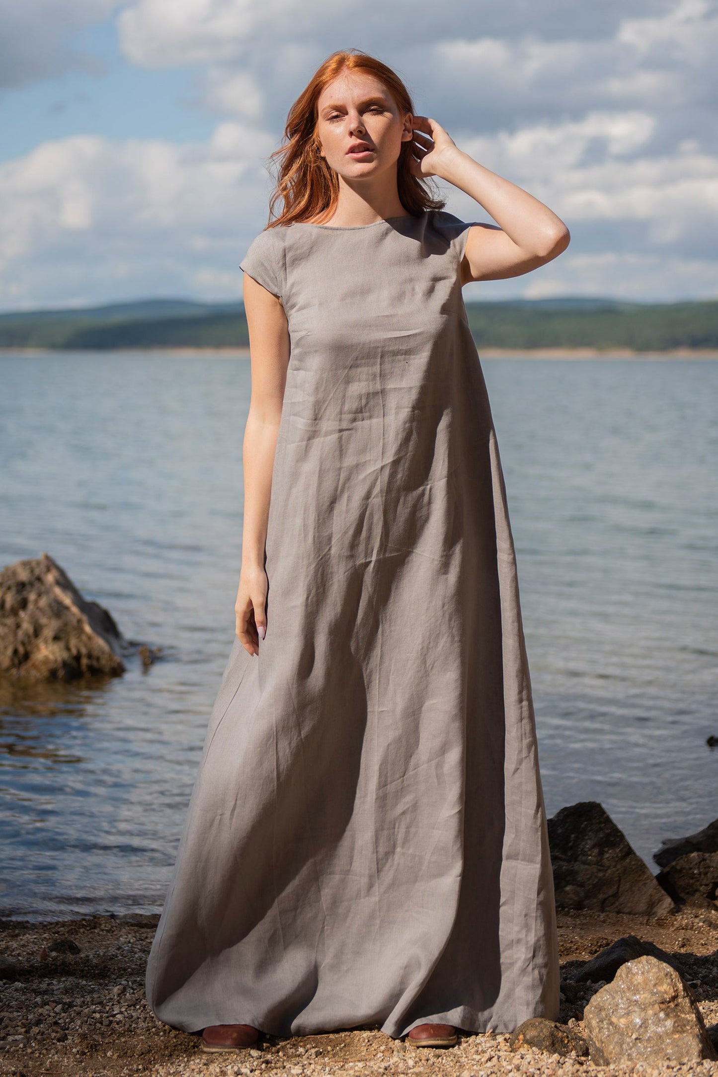 Woman wearing gray linen maxi dress with cap sleeves standing near water and rocks in natural light