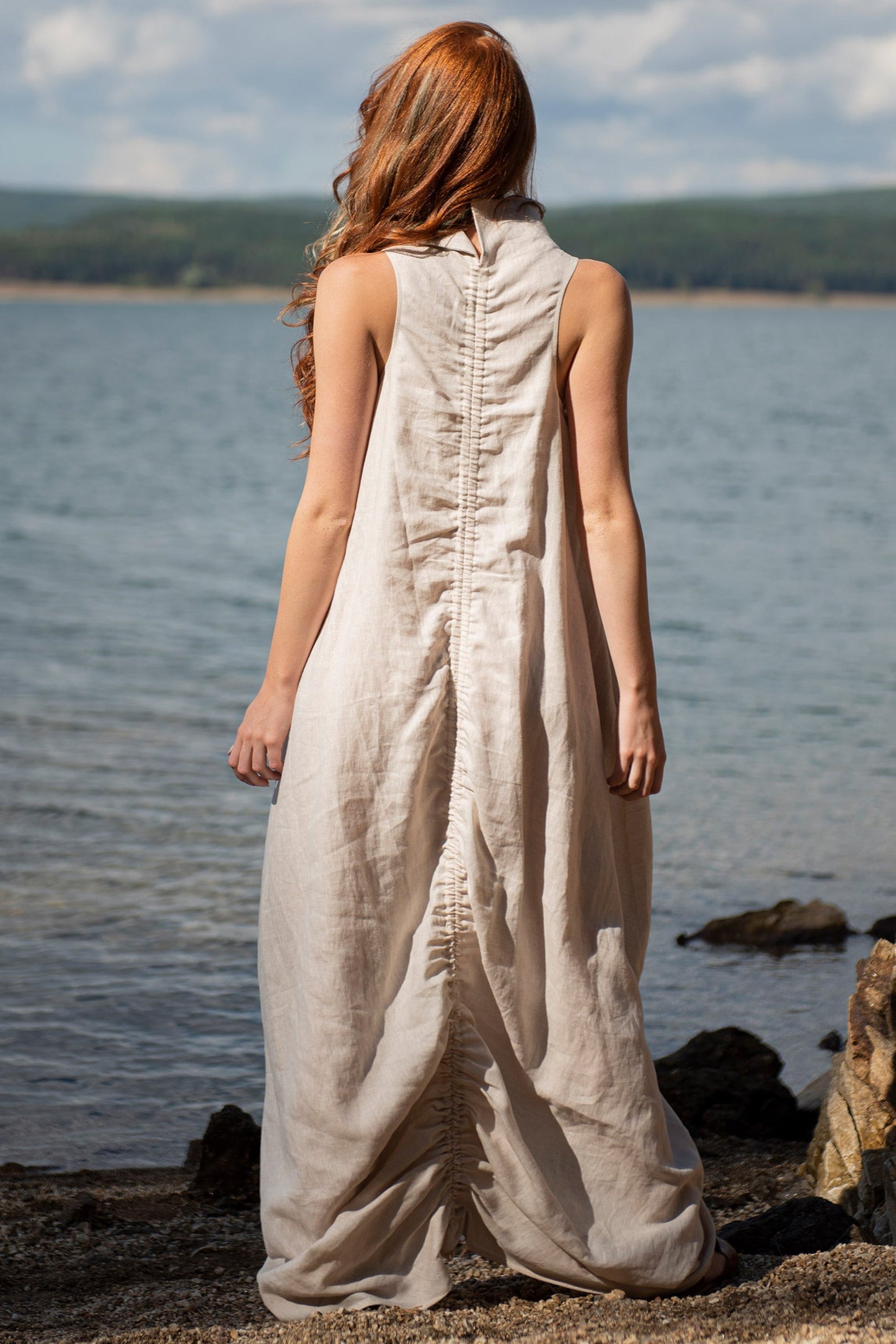 Back view of natural color cowl neck linen sleeveless maxi dress with ruched adjustable back worn by woman near water