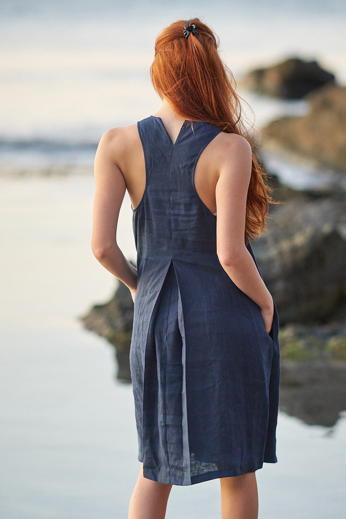 Woman wearing navy blue pinafore linen dress with halter neckline and pleated back detail standing by the water