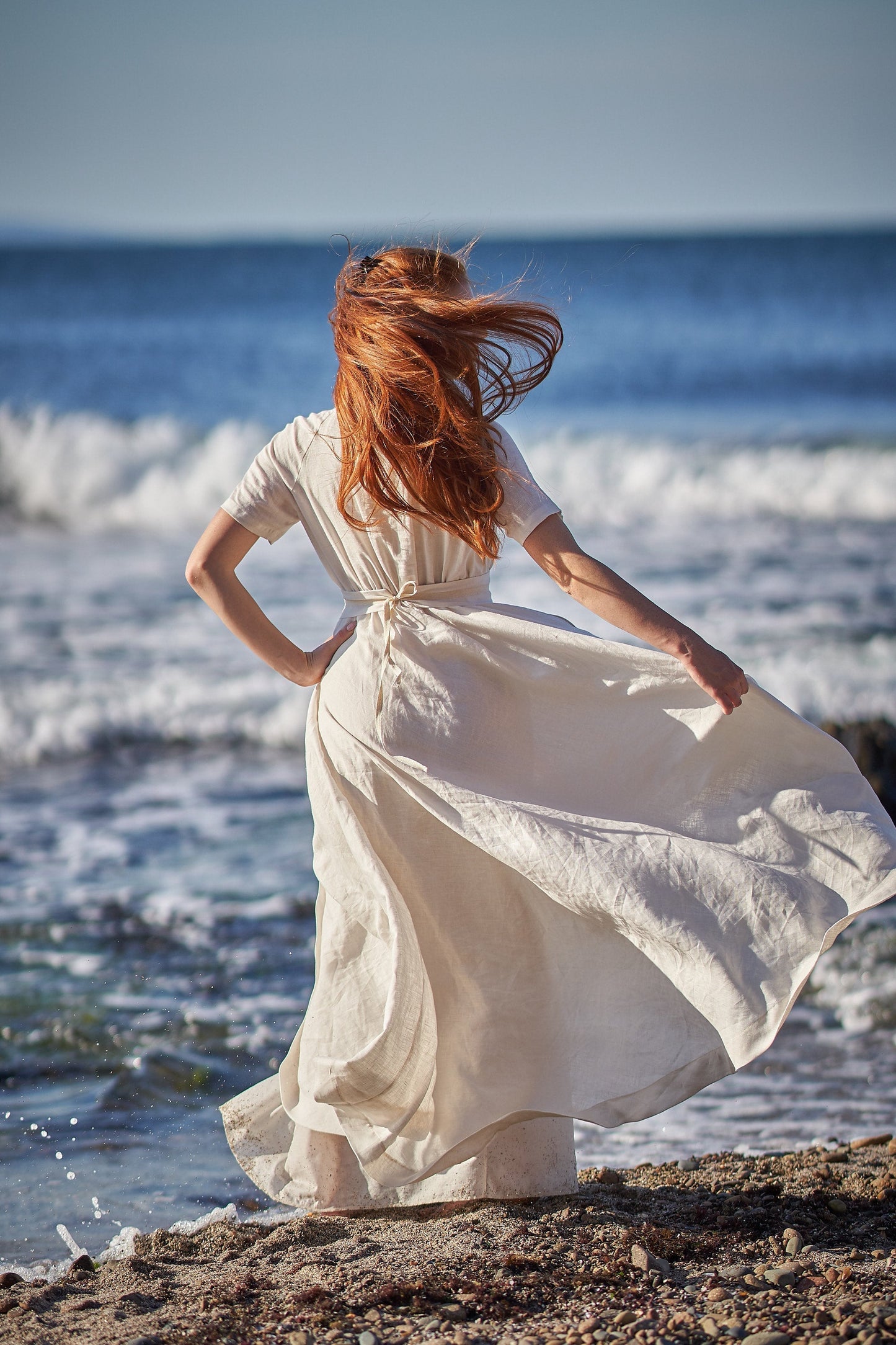 Woman wearing a creamy white summer linen wedding dress with short sleeves and wrap tie, standing on beach by the sea