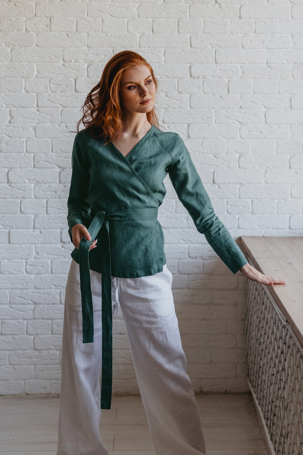Woman wearing a green top and white pants against a white brick wall.