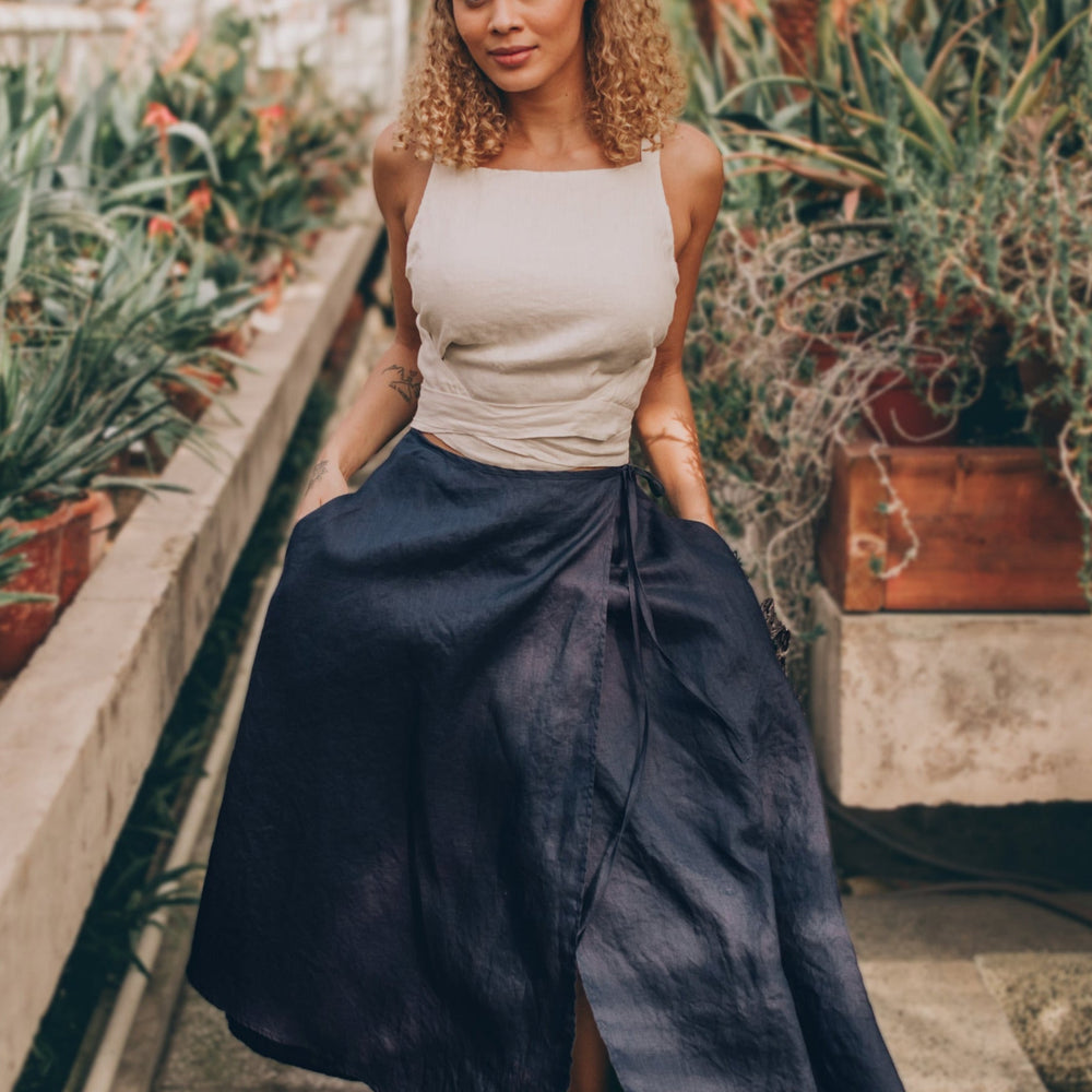 Midi length linen wrap skirt in Navy Blue with high rise and side pockets, modeled in a greenhouse.