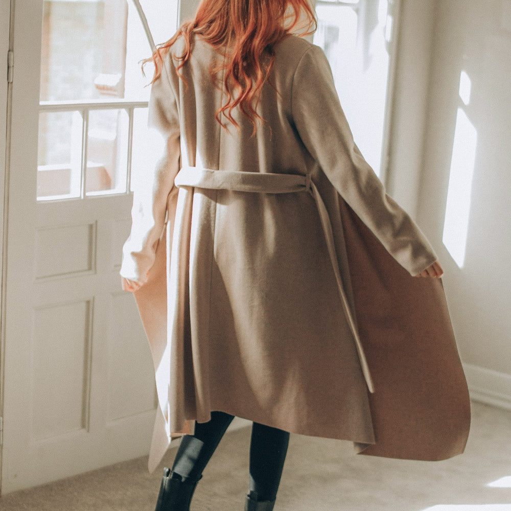 
                      
                        Woman wearing a beige coat standing in a sunlit room with large windows.
                      
                    