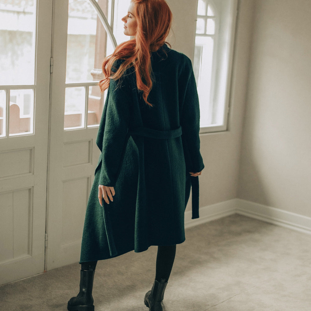 
                      
                        Woman with red hair wearing a green coat standing in a room with large windows.
                      
                    