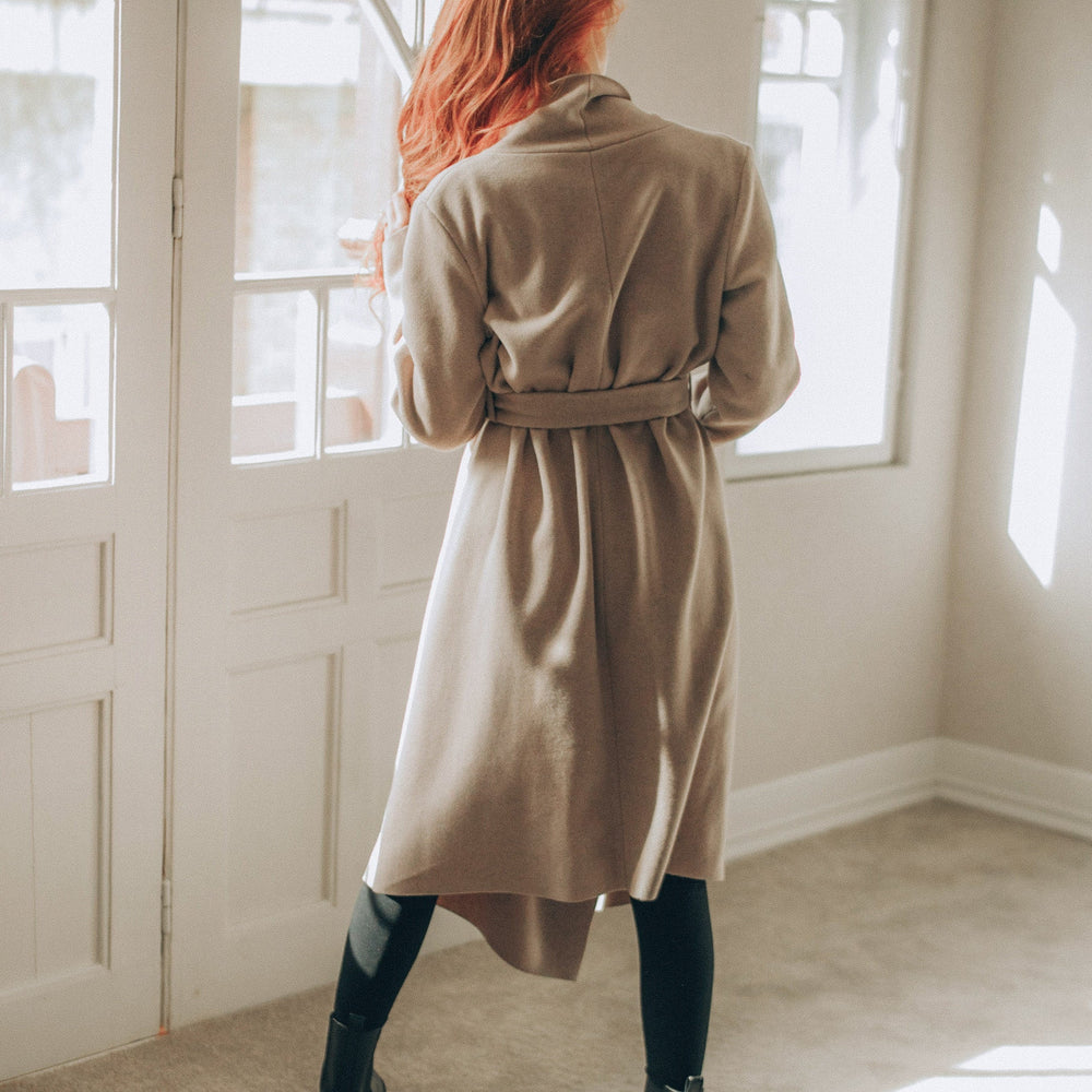 
                      
                        Woman wearing a beige coat standing in a sunlit room with large windows.
                      
                    