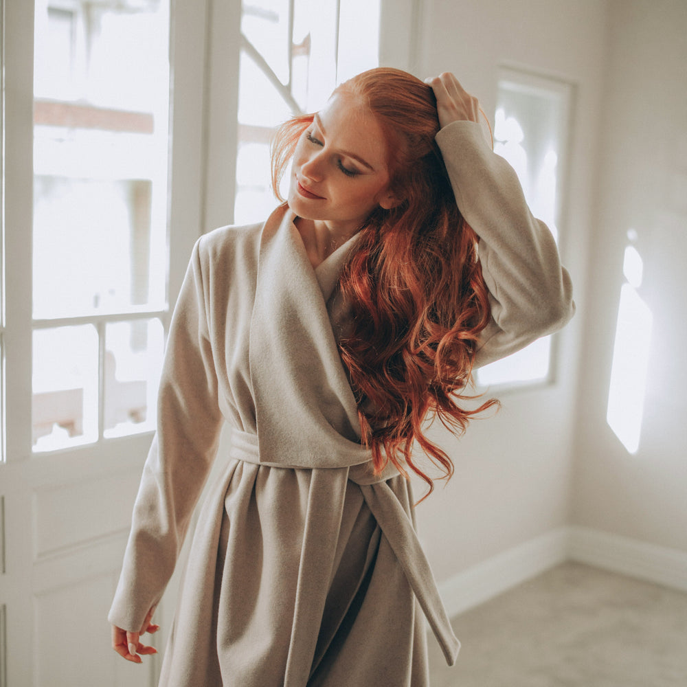 
                      
                        Woman with red hair standing in a sunlit room wearing a beige coat.
                      
                    