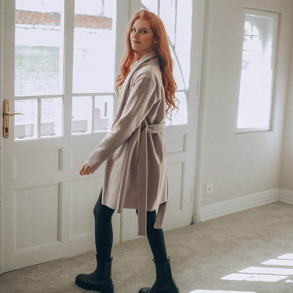 
                      
                        Woman in a beige wool short coat standing in a room with large windows.
                      
                    