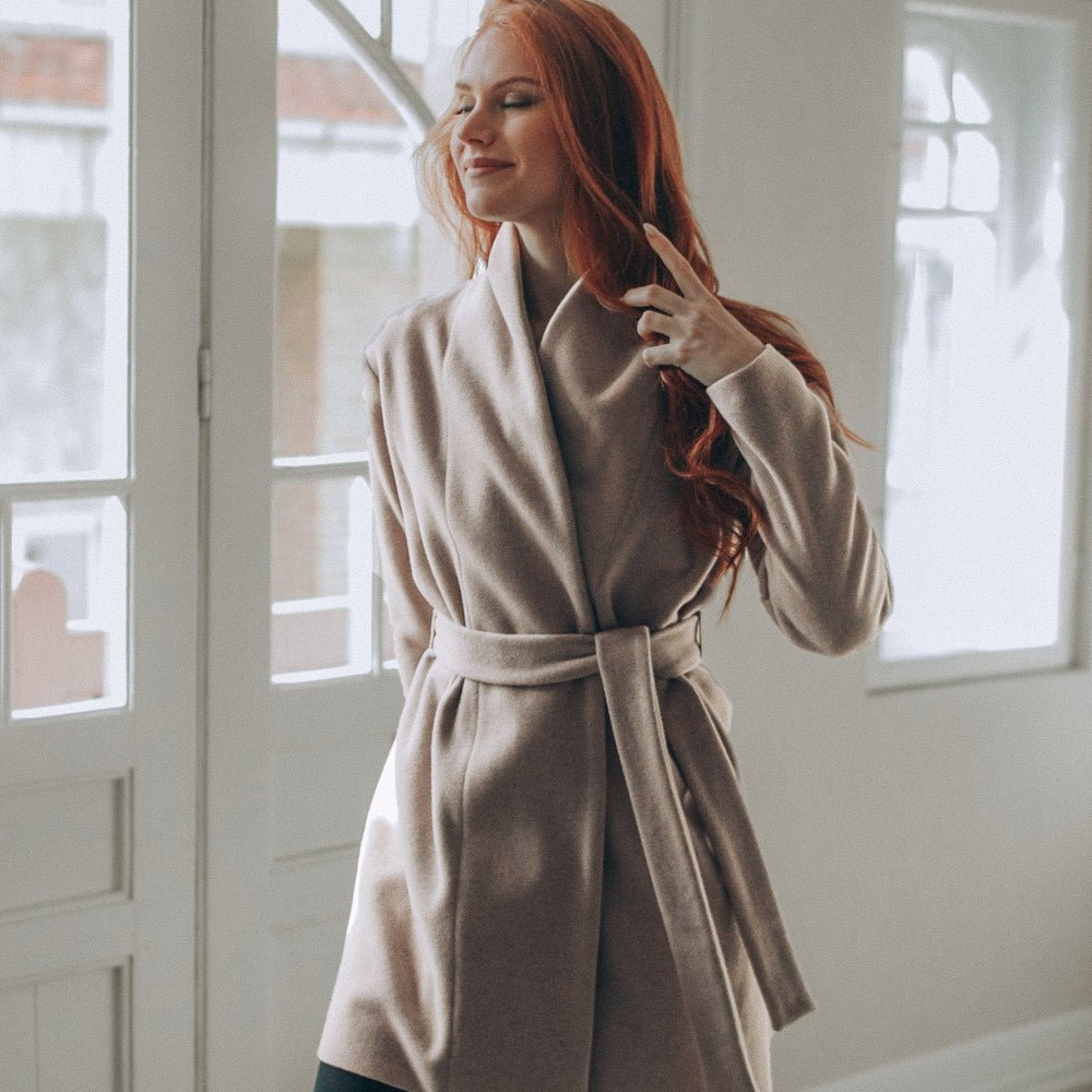 Woman wearing powder beige short wool cardigan coat with belt indoors near window