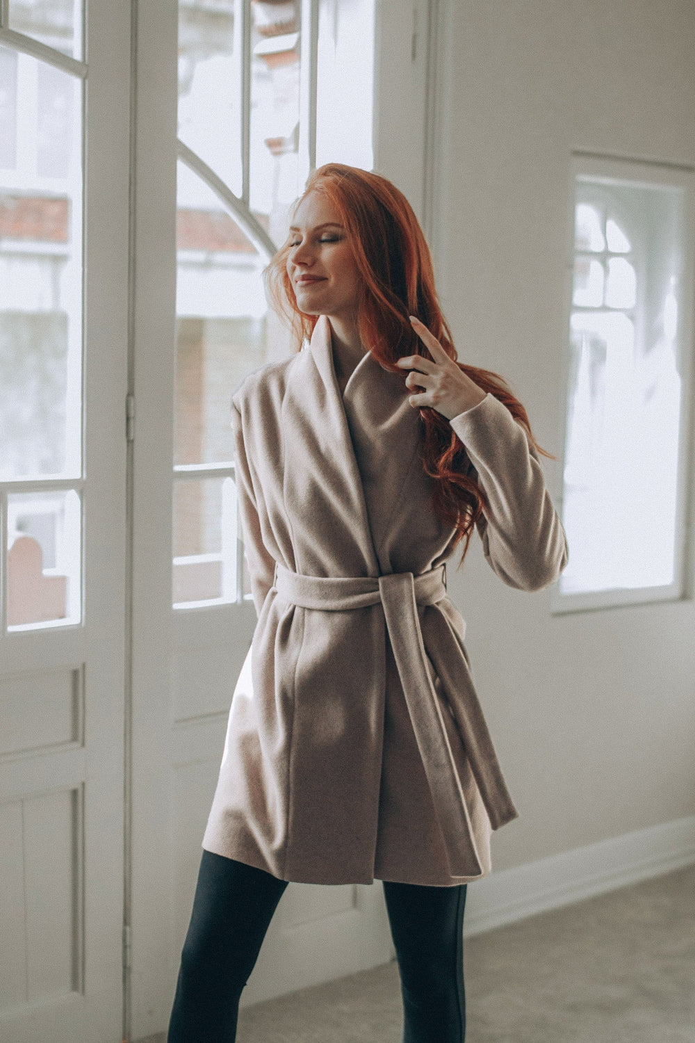 Woman wearing powder beige short wool cardigan coat with belt indoors near window