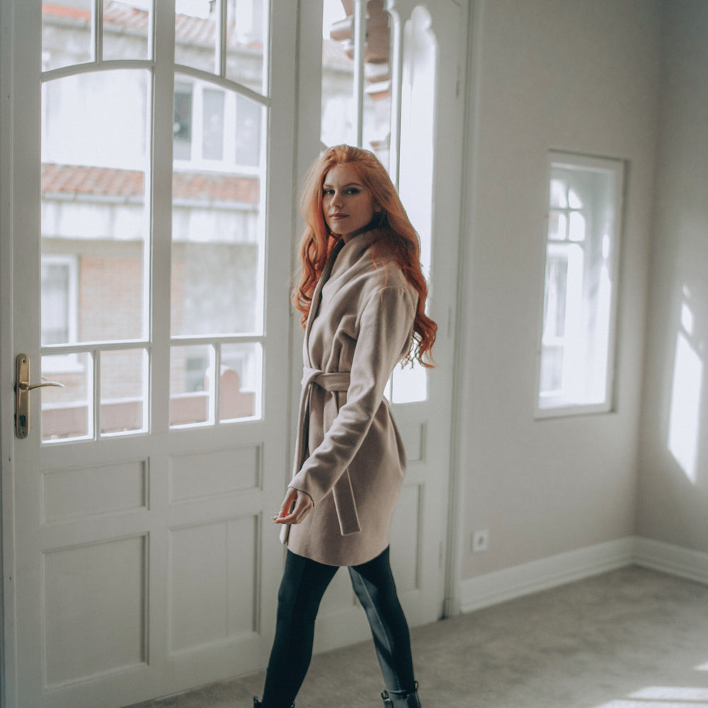 
                      
                        Woman in a beige wool short coat standing in a room with large windows.
                      
                    