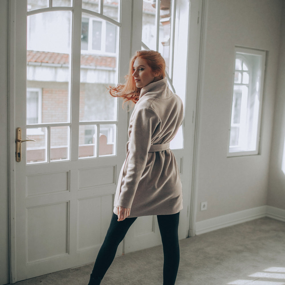 
                      
                        Woman in a beige wool short coat standing in a room with large windows.
                      
                    