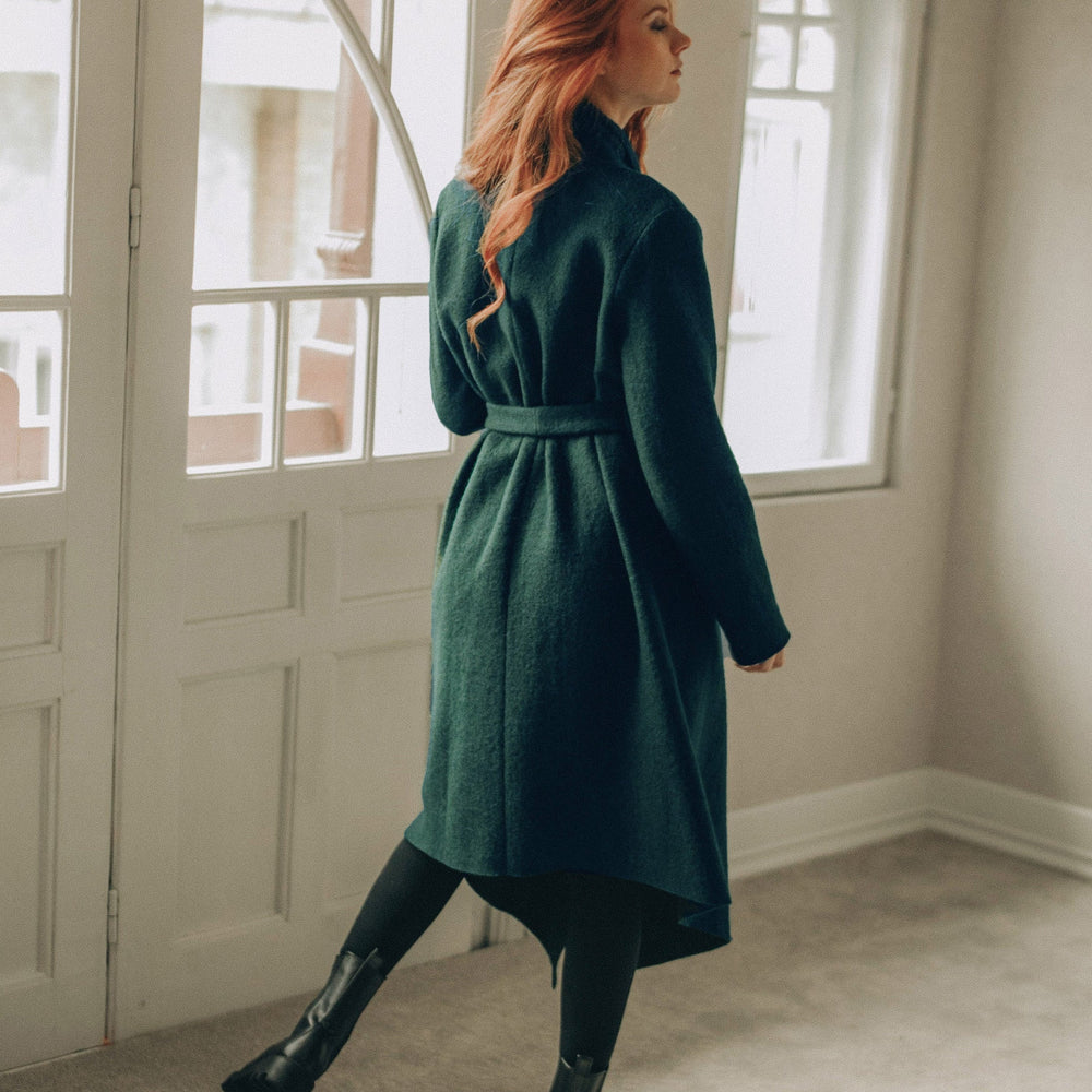 
                      
                        Woman in a green coat standing in a room with large windows.
                      
                    