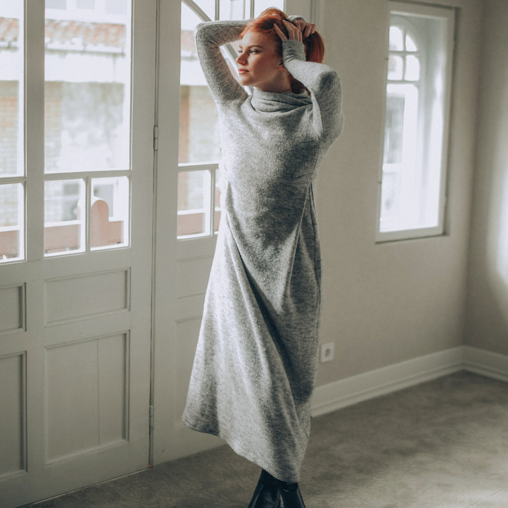 Woman in a long gray knitted dress standing in a room with large windows.