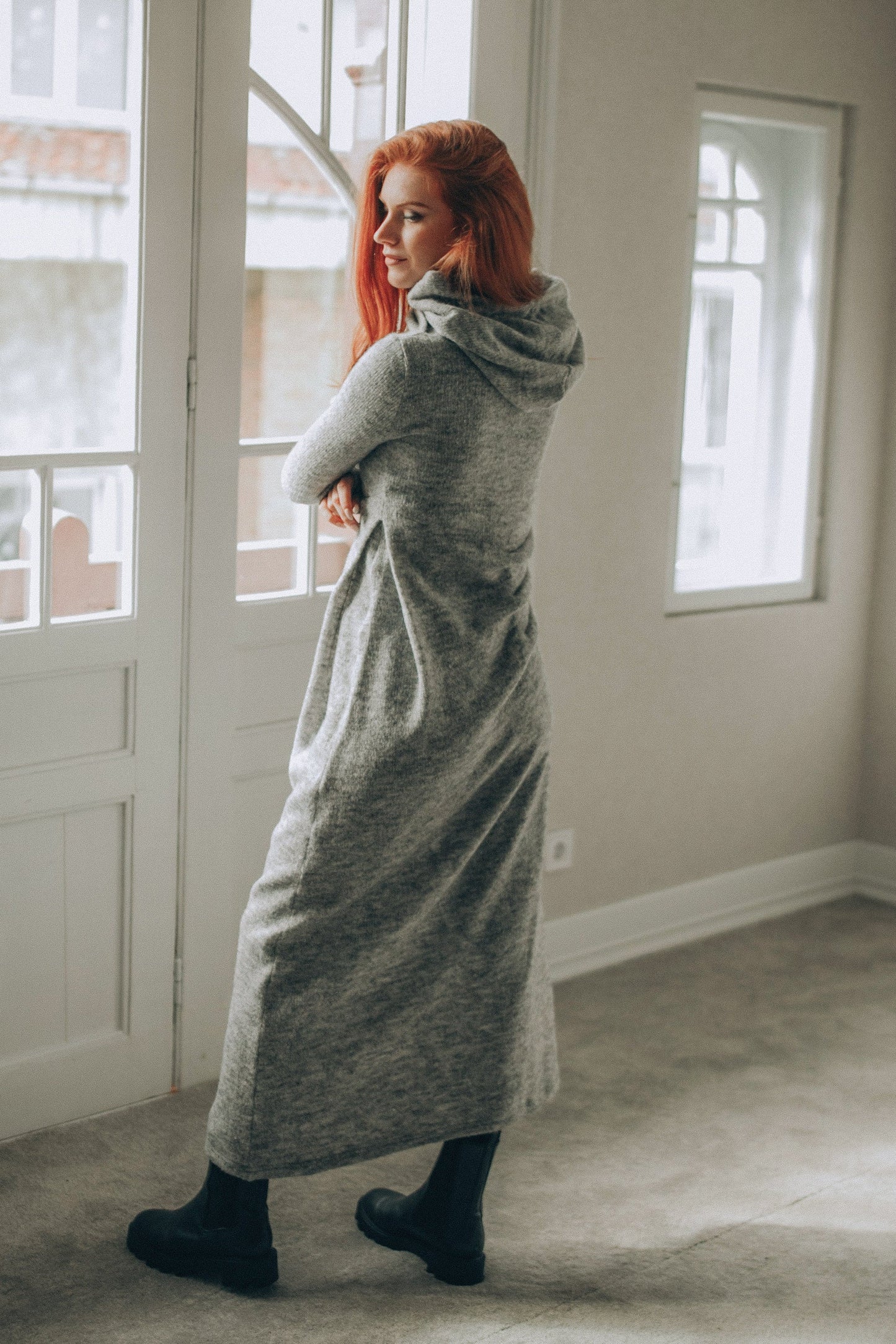 Woman wearing gray hooded knit dress with gathered side accent and black boots indoors