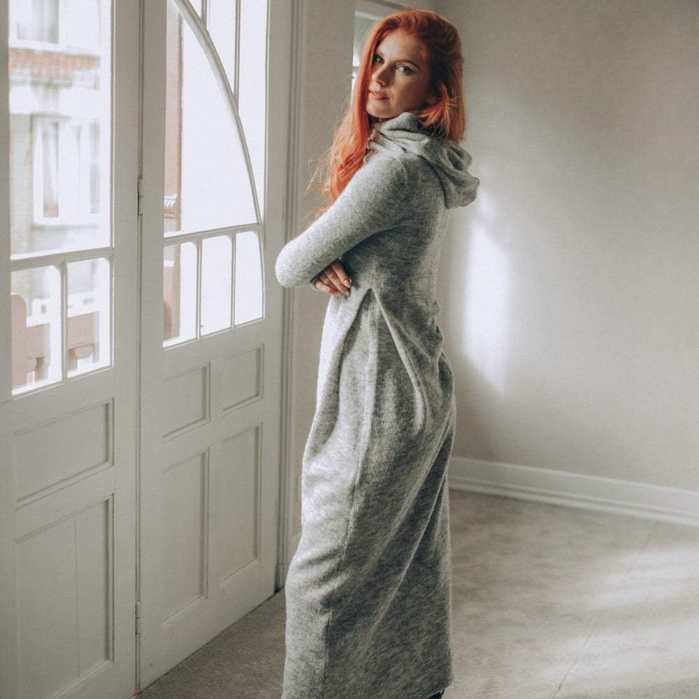 
                      
                        Woman in a long gray knitted dress standing in a room with large windows.
                      
                    