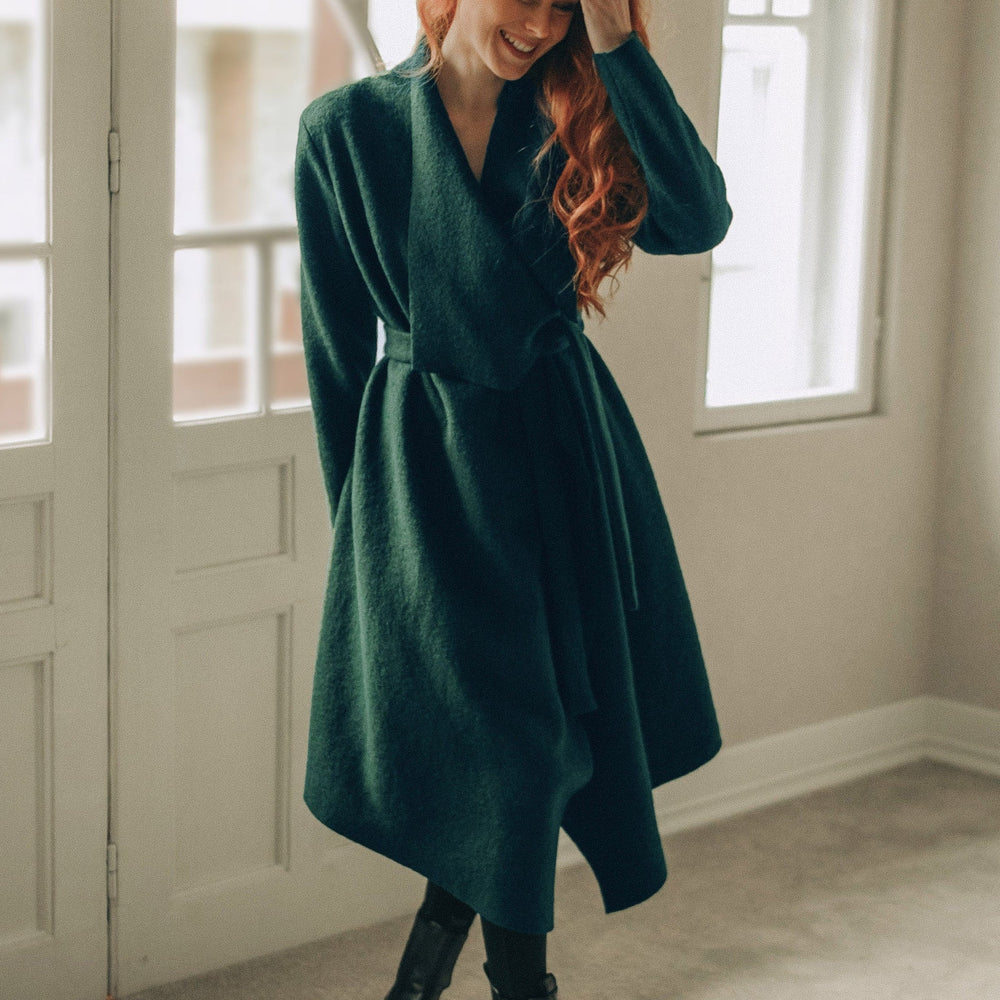 
                      
                        Woman in a green coat standing in a room with large windows.
                      
                    