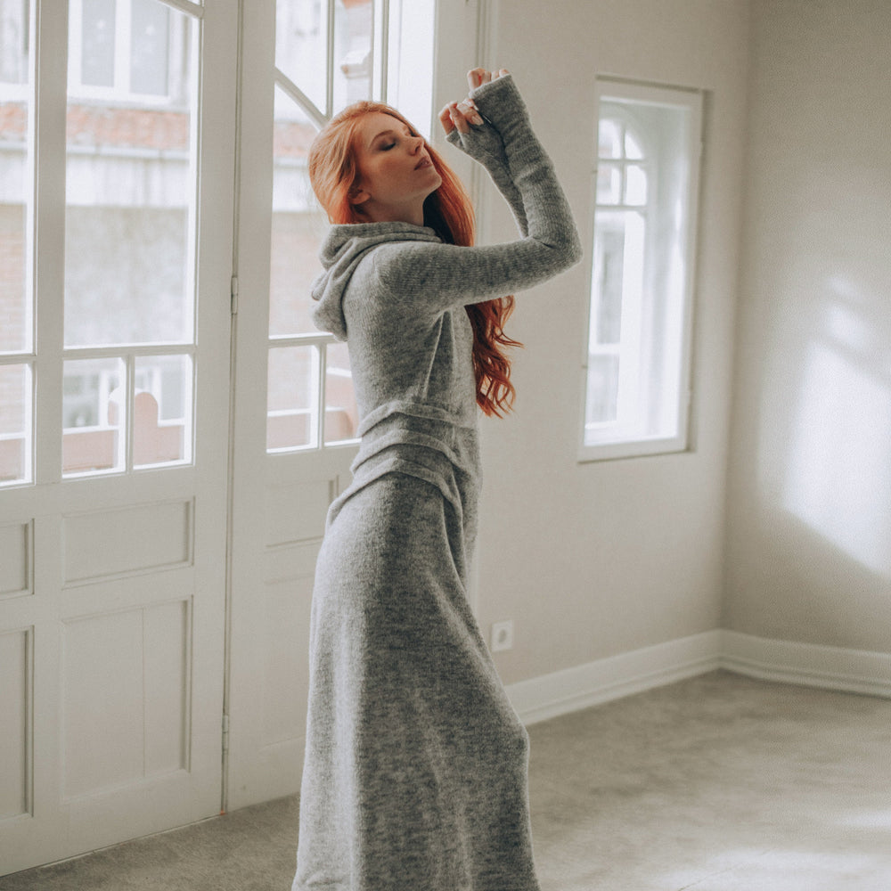 
                      
                        Woman in a long gray knitted dress standing in a room with large windows.
                      
                    
