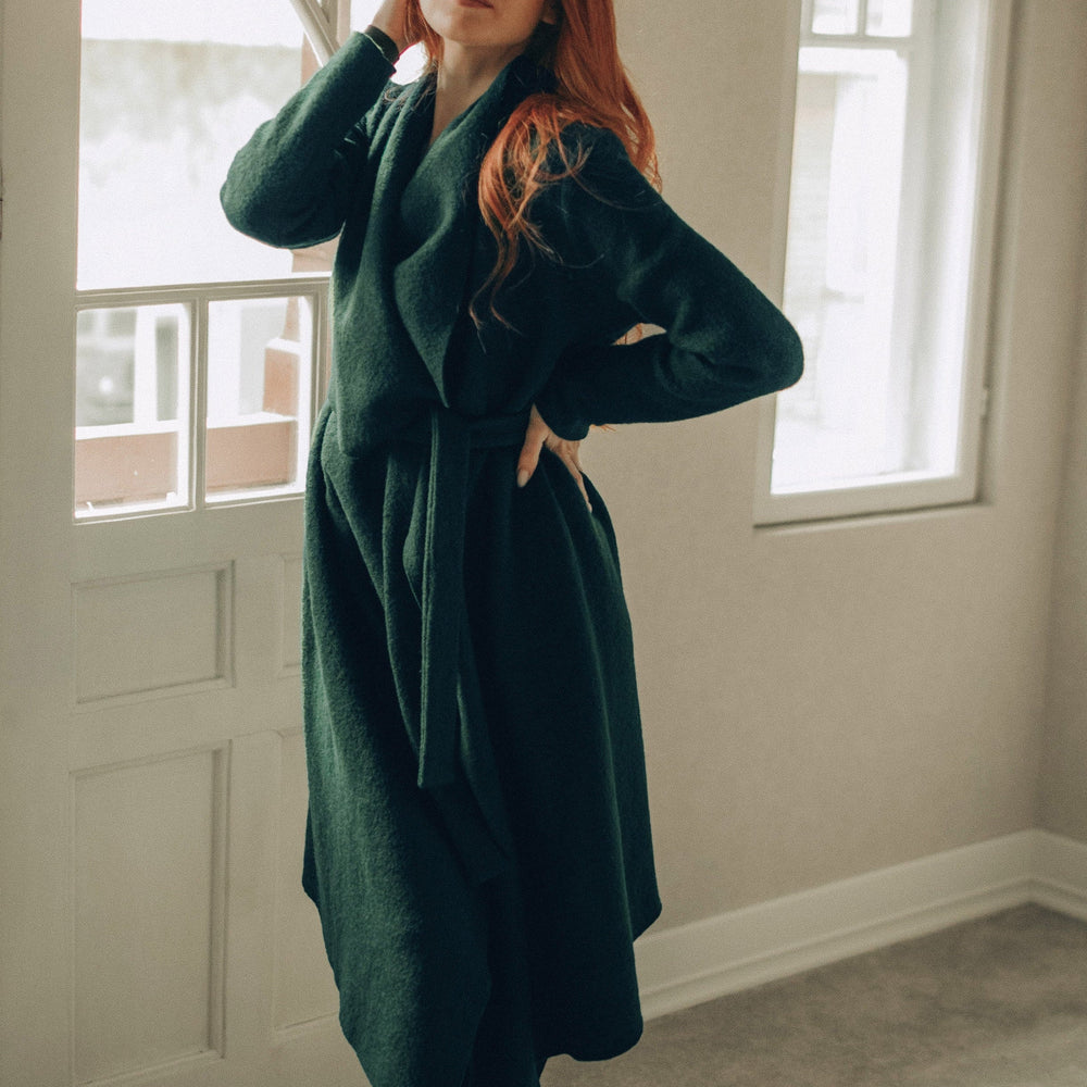 Person wearing a green coat standing in a room with large windows.