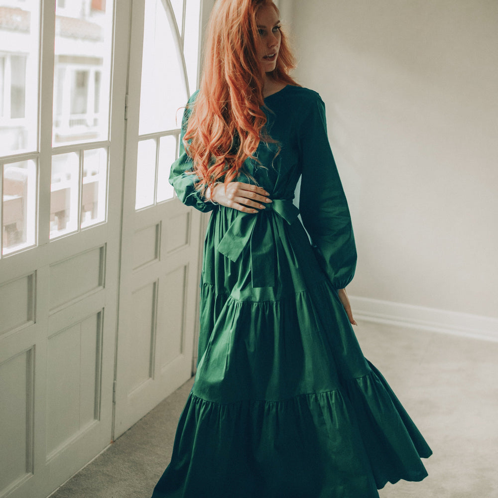 
                      
                        Woman in a green dress standing in a room with large windows.
                      
                    