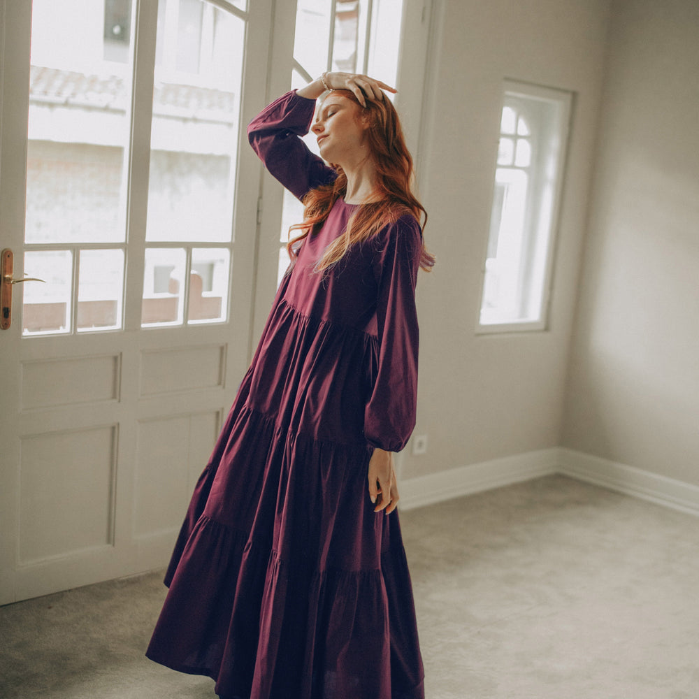Woman in a long burgundy dress standing in a room with large windows.