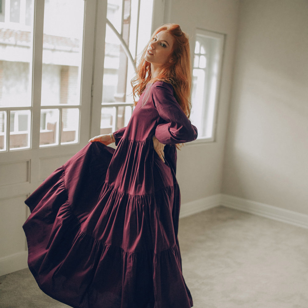 
                      
                        Woman in a long, dark purple dress standing in a room with large windows.
                      
                    